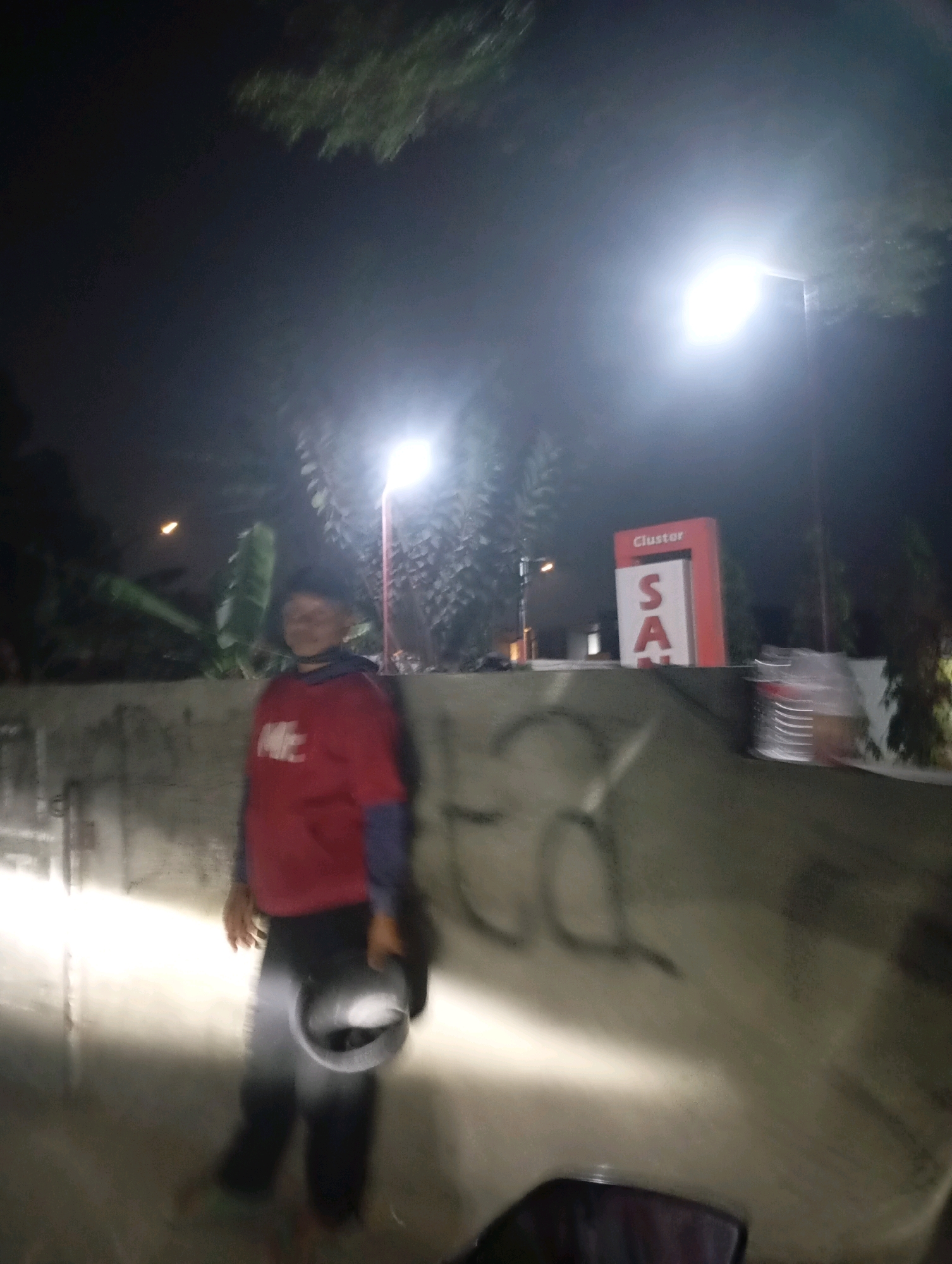 A person in a red sweatshirt and dark pants stands in front of a concrete wall at night. The person is holding a dark helmet in their right hand. The scene is lit by streetlights, one of which is visible in the upper right portion of the image. A sign with the word "Cluster" and the letters "S", "A", and what appears to be an "N" or "M" is visible in the background. The image appears to have been taken in Babelan, Indonesia.