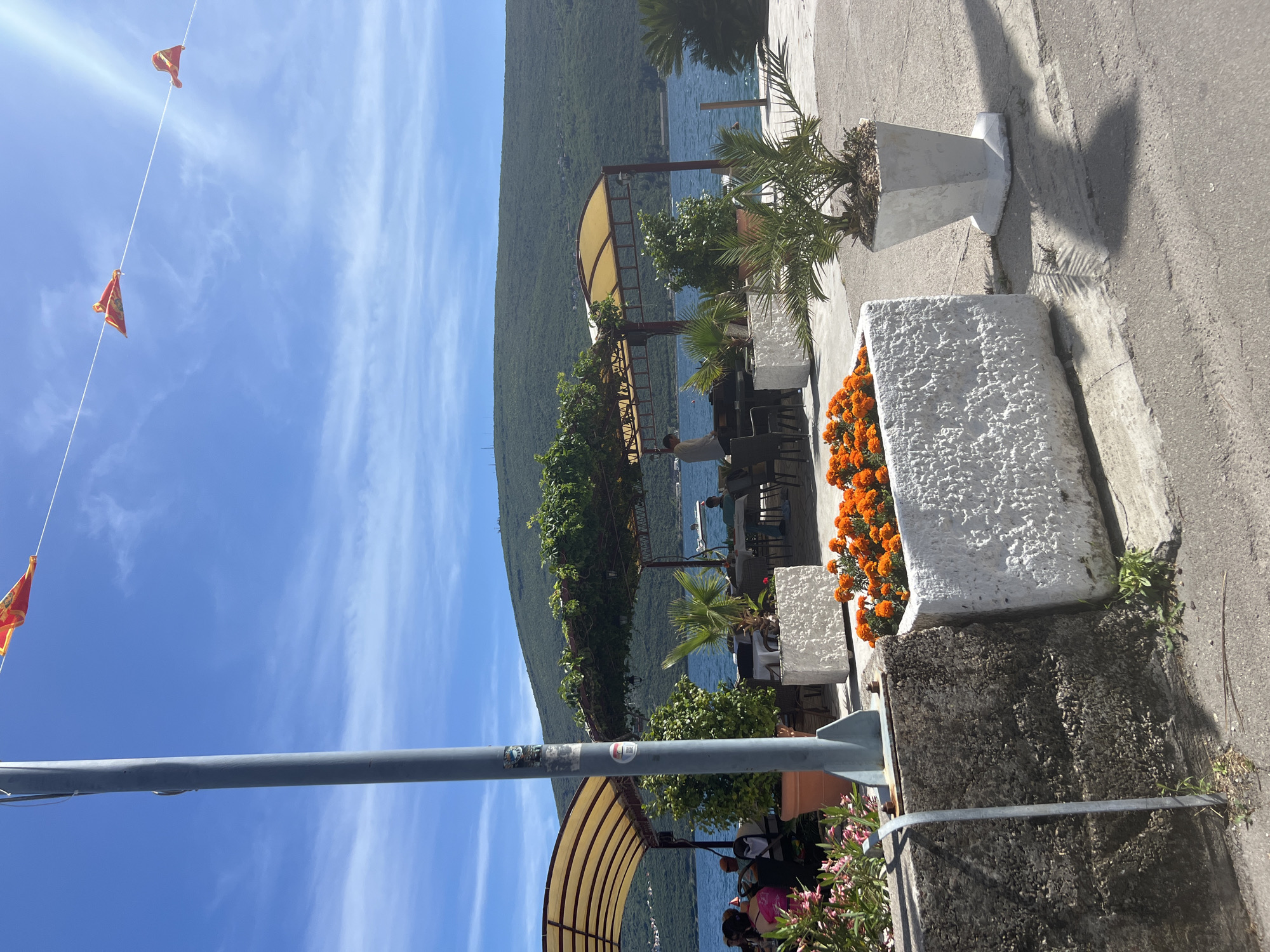 The image depicts a coastal scene in Đenovići, Montenegro. A view of the bay with a large, forested mountain backdrop is prominent. In the foreground, a roadside cafe or restaurant is visible with tables and chairs under yellow striped awnings. Planters with orange flowers and small palm trees line the sidewalk. A grey metal pole stands to the left of the frame with wires and small red triangular flags strung above it. People are seated at the tables of the cafe. The sky is blue with wispy clouds.