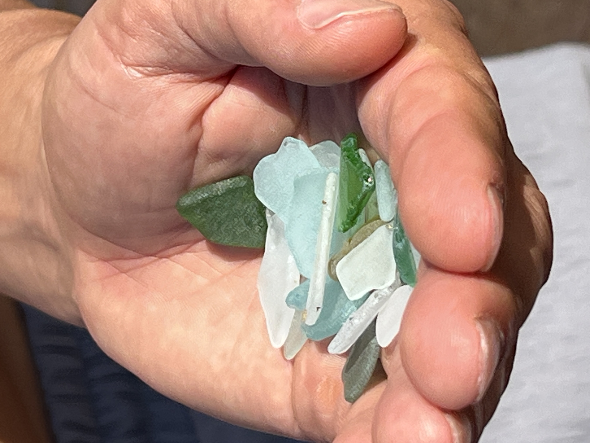 A close-up, overhead shot shows a person's cupped hand filled with sea glass. The hand, with its visible wrinkles and lifelines, is a light peach color. The sea glass pieces are varied in size and shape, with many appearing smooth and frosted from the sea. The colors range from deep green and aqua to clear white and light blue. The background is blurred, showing a dark blue surface and a white garment.