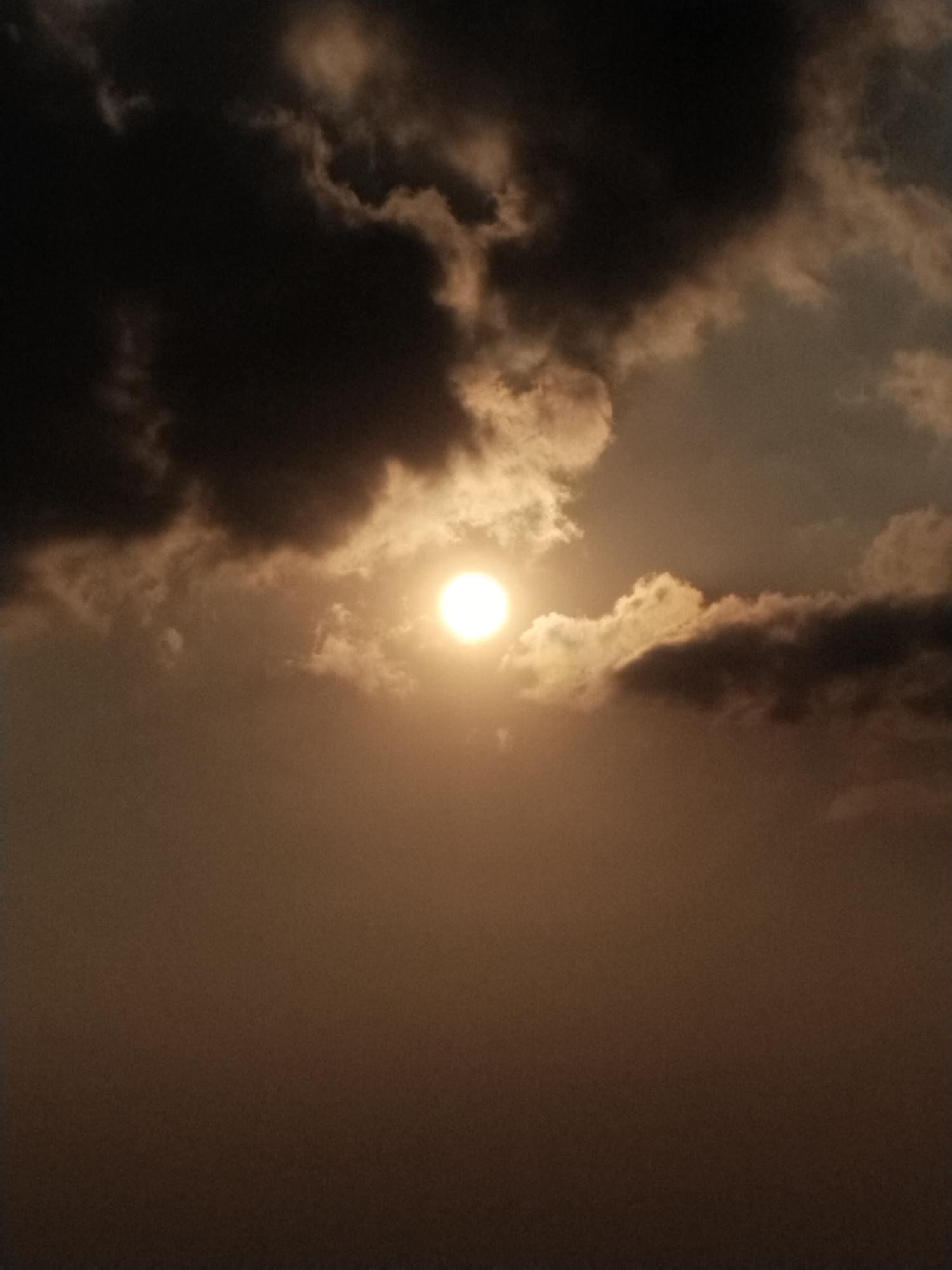 The image shows a cloudy sky with the sun partially obscured by dark clouds. The sun appears as a bright, glowing orb. The lower part of the image is a hazy, gradient of brown, suggesting a thick atmosphere or haze. The scene is an outdoor environment, likely a sky view, with no specific urban or natural landmarks to identify a precise location. There are no people, objects, or actions depicted in the image.