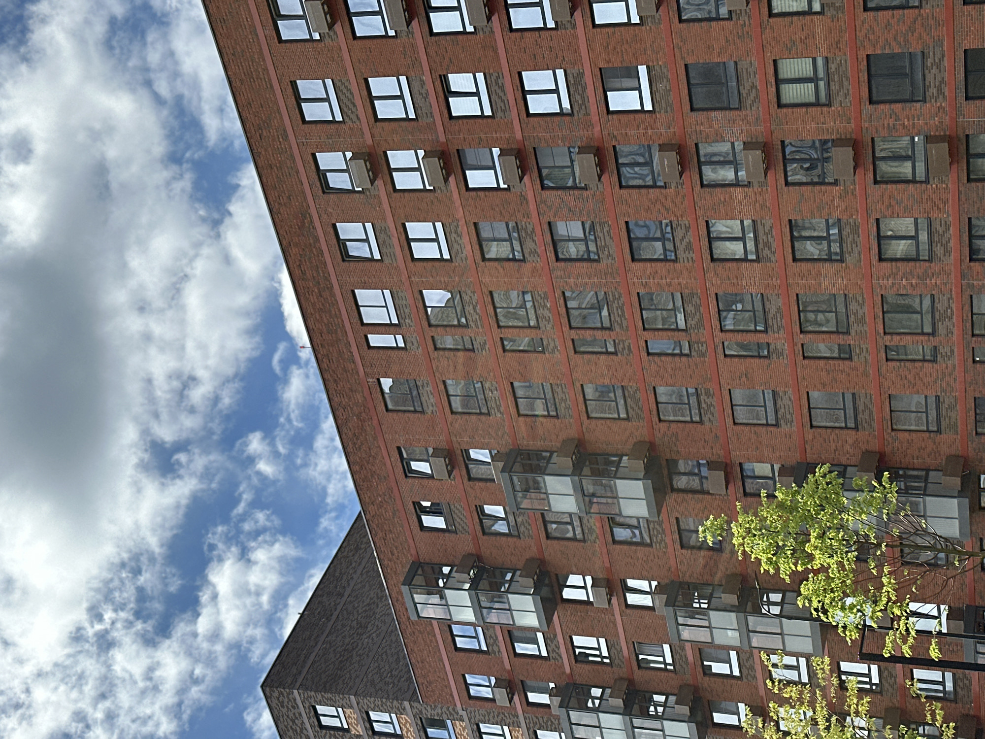 A close-up, eye-level, vertical shot showcases the facade of a modern apartment building constructed from red brick in Znamya Oktyabrya, Russia. The building features numerous windows with dark frames, some of which are clear, reflecting the sky and clouds, while others appear tinted or opaque. Small, rectangular air conditioning units are visible beneath many of the windows. Several cantilevered glass balconies, enclosed with metal frames, are interspersed throughout the building's levels. A darker, presumably older, building section is partially visible on the left side of the frame, with a few windows. The sky above is a dynamic mix of blue and white clouds, with some appearing more dense and overcast. In the foreground, the vibrant green foliage of a young tree with slender branches partially obscures the lower portion of the building, suggesting a residential or urban setting. The image captures the architectural details of the apartment building against a cloudy sky.