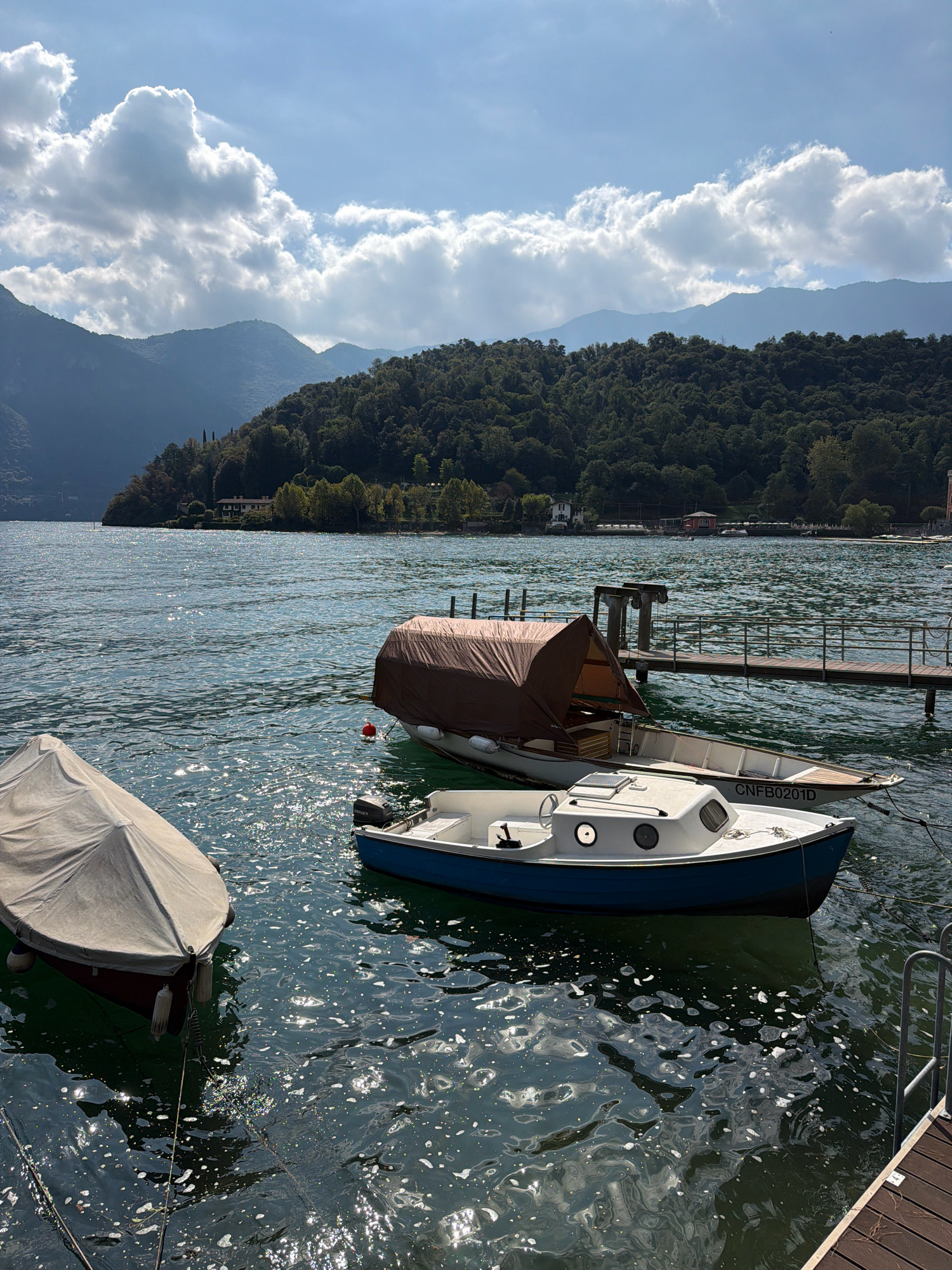 The image depicts a scenic view of Lake Como in Tremezzina, Italy. The water is choppy and reflects the sunlight, creating a shimmering effect. Several boats are moored in the foreground and middle ground. One boat on the left is covered with a light-colored tarp. In the center, a larger boat is partially covered with a brown tarp, and a smaller blue and white boat is in front of it. A wooden pier extends into the water from the right side of the frame. In the background, lush green hills covered in trees rise up, with a few buildings visible nestled among the foliage. The sky is partly cloudy, with large white clouds contrasting against a blue sky. The overall impression is one of a tranquil, sun-drenched day on the lake.