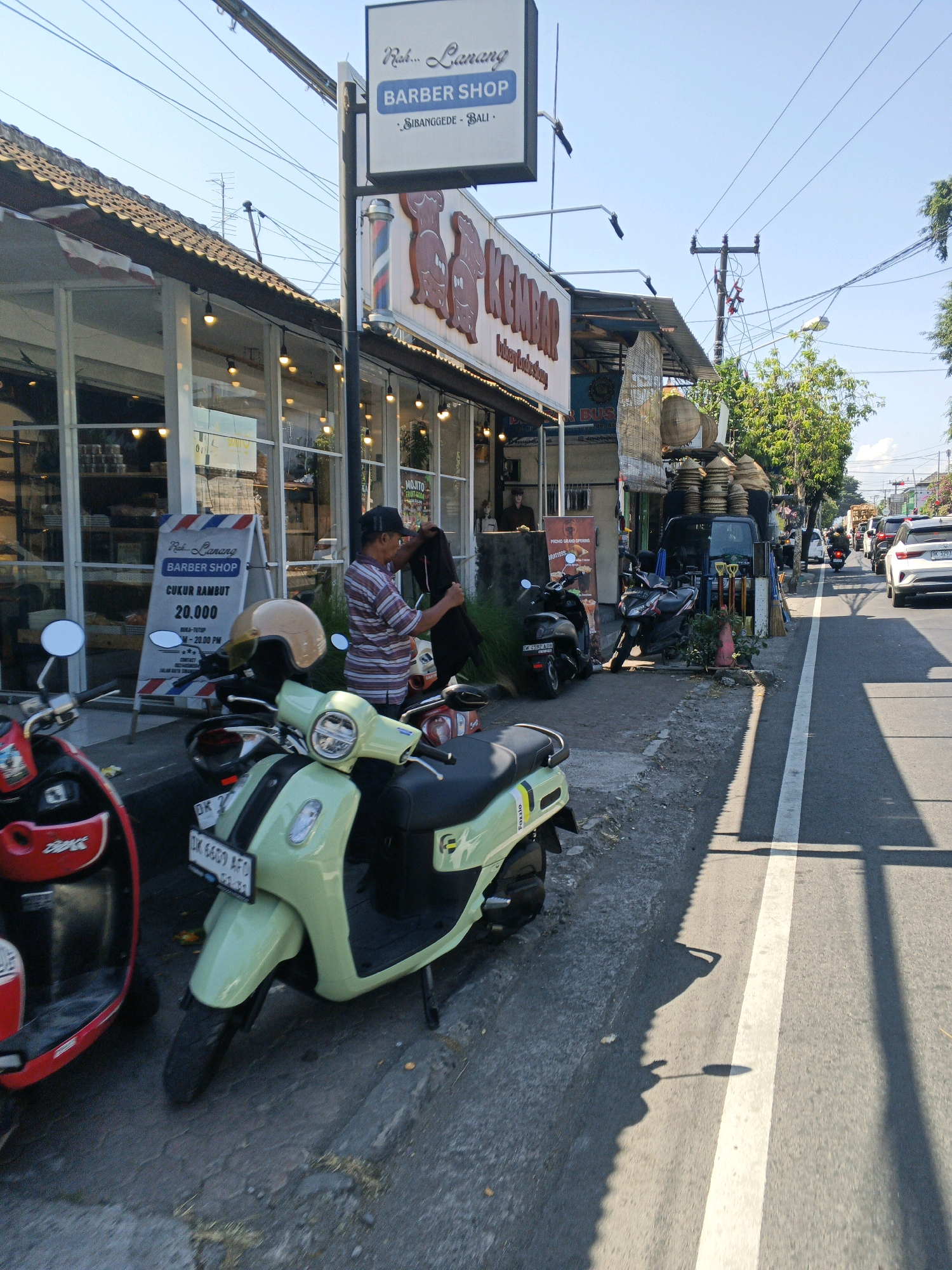 The image captures a street scene in Abiansemal, Indonesia, on a clear, sunny day. In the foreground, two scooters are parked on a sidewalk next to a road. A light green scooter is positioned centrally, while a red scooter is partially visible on the left. Behind the scooters is a barber shop named "Ruk... Lanang Barber Shop," indicated by a sign with text and a barber pole graphic. The shop has large glass windows, with some visible interior shelves and lighting. A man wearing a striped shirt and a cap stands near the shop entrance, holding a dark garment. Further down the street, more parked vehicles, including another scooter and a black van, are visible. The road features a white dividing line. In the background, trees line the street, and utility poles with numerous wires are present. Traffic, including a white car and a motorcycle, is moving along the road. The overall environment suggests a commercial area with a relaxed, daytime atmosphere.