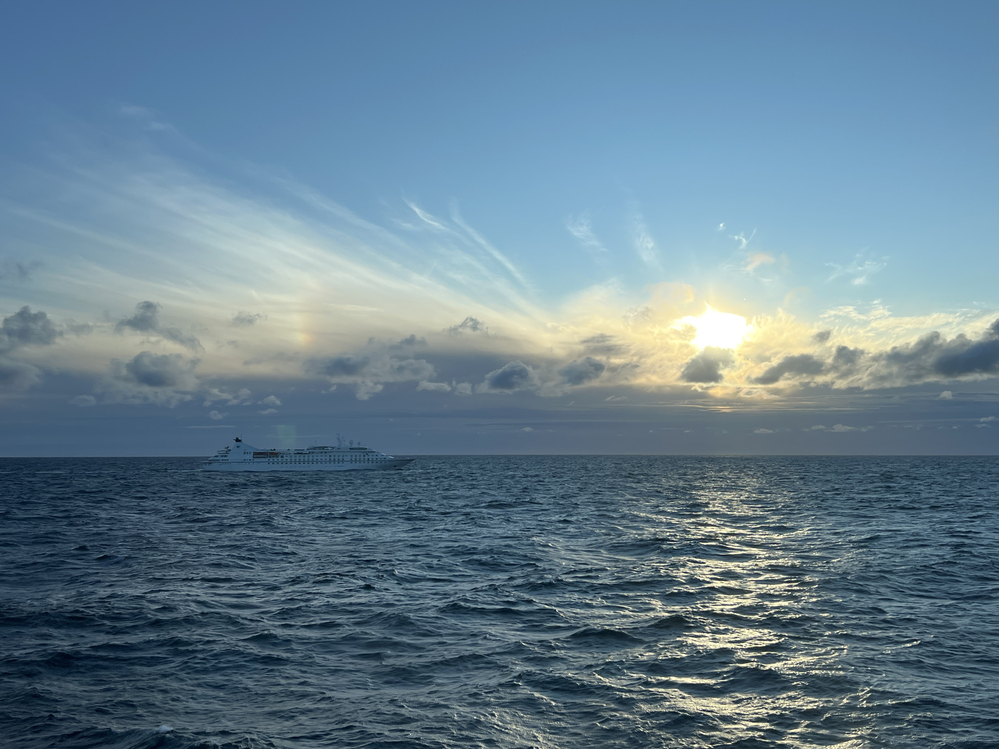 An expansive view of the sea shows a white cruise ship sailing on the horizon. The sky above is a clear blue, with streaks of cirrus clouds and a bright sun partially obscured by darker, more scattered clouds. The sun's rays create a shimmering reflection on the choppy, dark blue water. The overall scene conveys a sense of calm and vastness. The location is noted as Unknown, Poland.