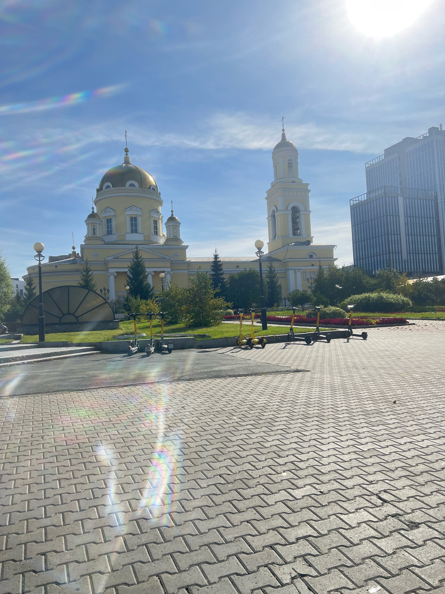 The image captures a sunny day scene in Yekaterinburg, Russia, likely in front of the Church of All Saints. The foreground features a paved walkway with a distinctive interlocking brick pattern. Scattered across this walkway are several yellow electric scooters parked in a row. In the middle ground, a large, yellow, domed church with a tall bell tower stands prominently. The church is flanked by green trees and bushes. Behind the church, a modern, glass-fronted skyscraper rises into the bright blue sky, which is partly obscured by thin, wispy clouds and a noticeable lens flare from the sun. The overall environment suggests an urban setting where historical architecture meets contemporary development.