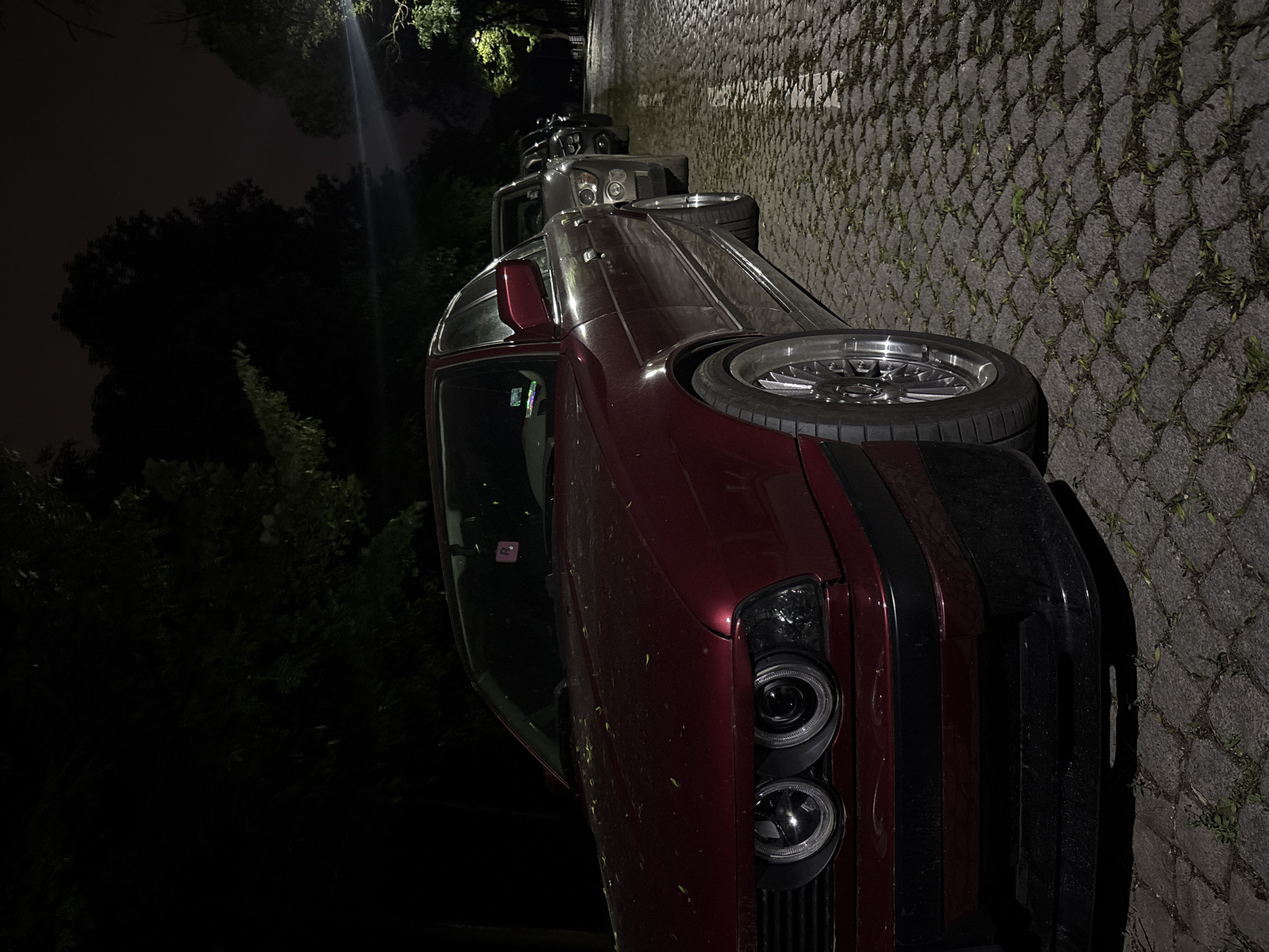 The image shows a maroon car parked on a cobblestone road in Sofia, Bulgaria at night. Other cars are parked behind it, illuminated by streetlights. The surrounding area appears to be a park or green space.