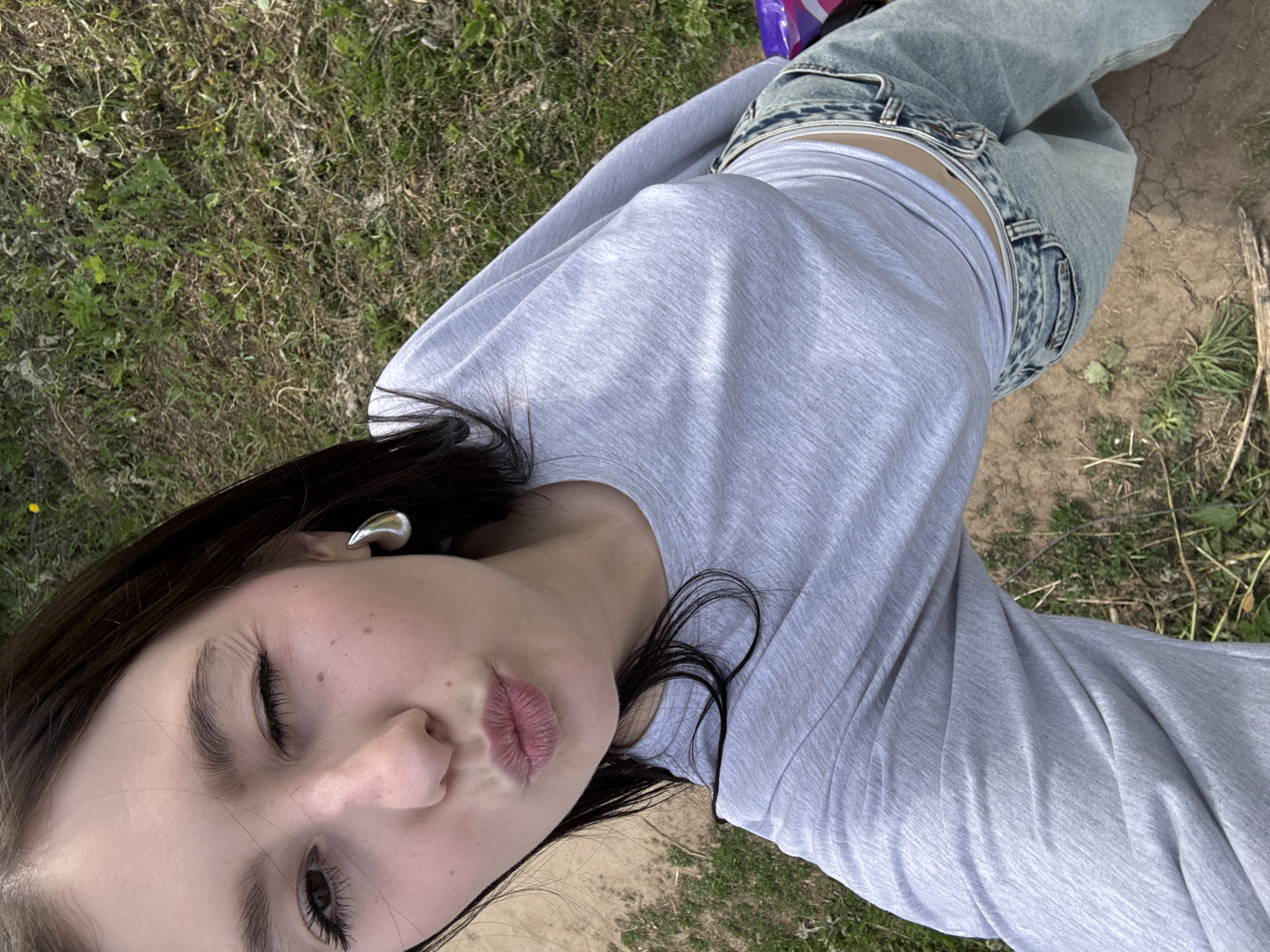 A young woman is positioned outside, lying on a grassy area adjacent to a patch of dirt. She is wearing a light grey long-sleeved shirt and faded blue jeans, which are unbuttoned revealing some of her midriff. She is wearing a silver earring. Her eyes are partially closed. Her lips are pursed. The location, Фоки, Russia, is not discernible from the visual information provided.