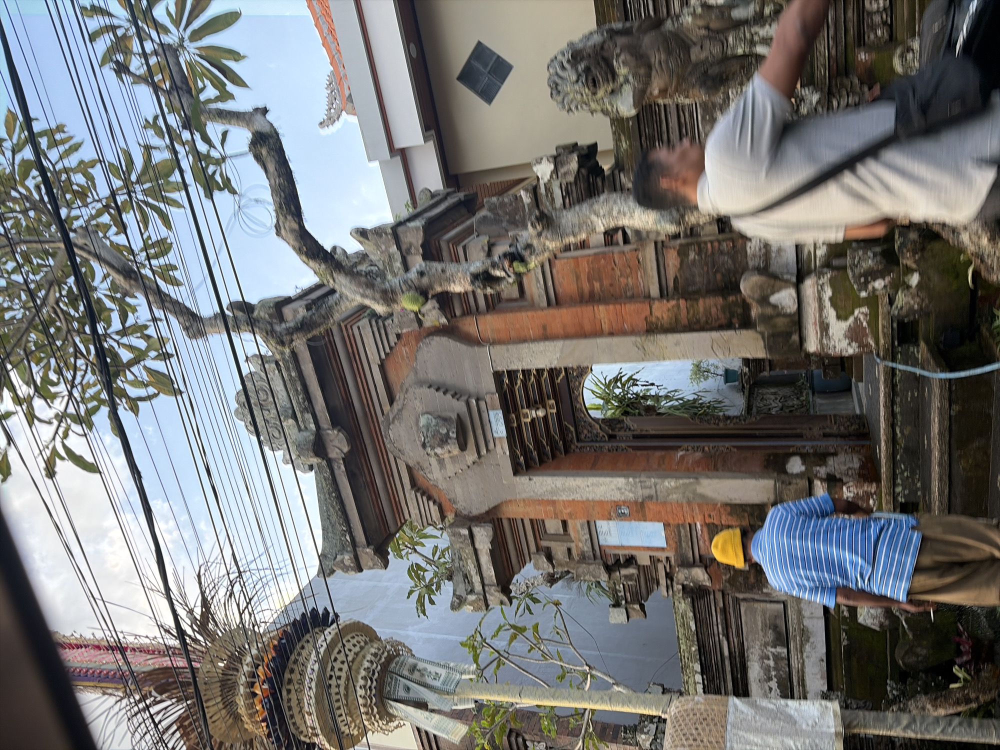 The image shows a street scene in Ubud District, Indonesia. A man wearing a yellow hat and a blue and white striped shirt is walking away from the camera, holding a cigarette. He is walking up a set of stone stairs in front of an ornate brick gateway. To the right of the gateway, there is a stone sculpture of a mythical creature. Another man, wearing a grey shirt and a black bag slung across his shoulder, is partially visible in the foreground, blurred by motion. Above the gateway, a tree with gnarled branches stretches towards the sky, and overhead power lines are strung across the frame. In the background, a building with a red-tiled roof is visible. The scene appears to be a residential or temple area, characteristic of Ubud.