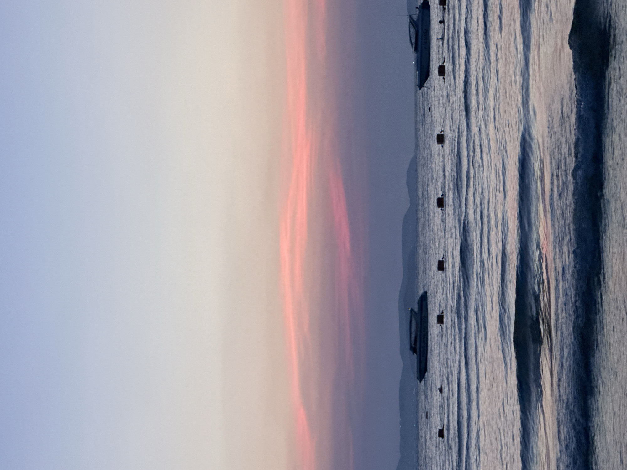 A vertical photograph displays a sunset over the ocean at Pattaya City, Thailand. The left side of the photograph features a gradient of colors: light blue, white, and pink in the sky. The right side depicts the ocean's surface, with wavy patterns and a few boats anchored with buoys. Dark silhouettes of distant hills can be seen near the horizon where the sea meets the sky.