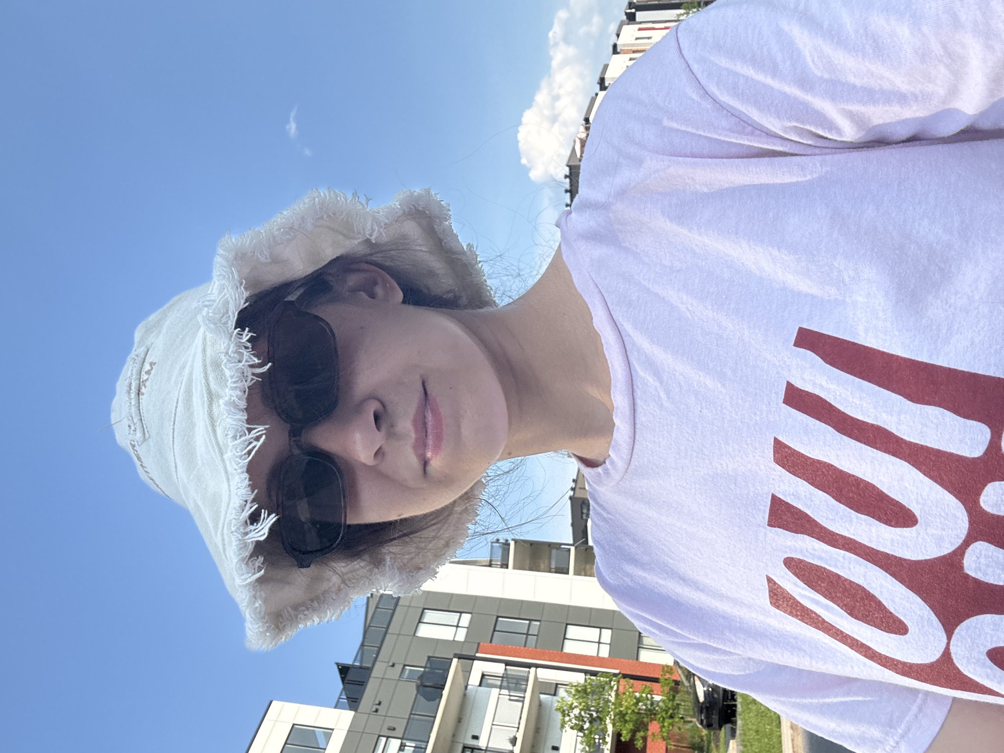 A young woman is taking a selfie outdoors in the city of Gatineau, Canada. She is wearing a light-colored bucket hat with frayed edges, dark sunglasses, and a light pink t-shirt with a large red graphic print. Behind her, a modern, multi-story building complex with balconies and balconies is visible, along with a bright blue sky and some clouds. A street and some greenery are also present in the background. The woman appears to be in good spirits, with a slight smile on her face. The overall scene suggests a casual, sunny day.