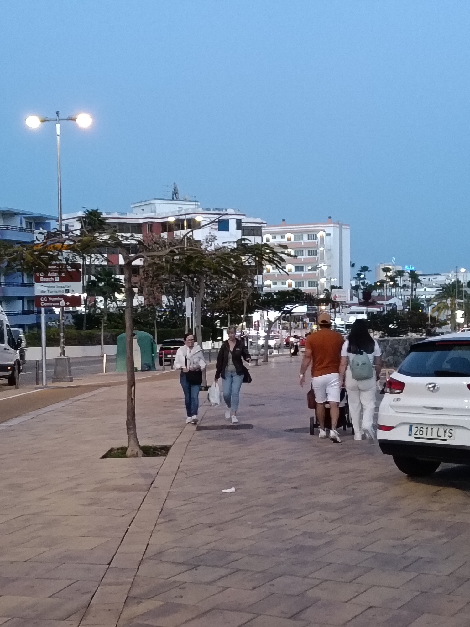 Captured at dusk in San Bartolomé de Tirajana, Spain, the scene unfolds along a brick-paved walkway. Two women walk together, one in a white jacket and jeans, the other in a dark jacket and jeans, both carrying white bags. A couple walks in the same direction, the man in an orange t-shirt and white shorts pushing a stroller. The woman is wearing white pants and a light green backpack. Parked on the right side of the frame is a white car with the license plate "2611 LYS." A sign directs towards the "CC Yumbo Centrum". Streetlights illuminate the scene, and buildings can be seen in the background.