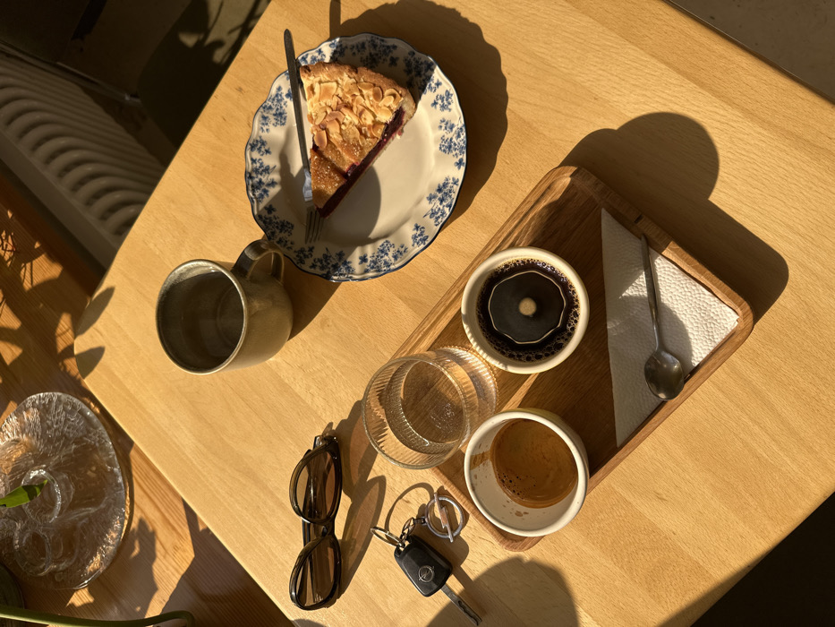 A high-angle photograph captures a cafe table scene in Kazan, Russia, illuminated by direct sunlight creating strong shadows. A light-colored wooden table serves as the primary surface. In the center, a wooden serving tray holds a small white ceramic cup with espresso, a clear ribbed glass filled with water, and another white ceramic cup containing dark coffee with a distinct circular pattern on its surface. A small metal spoon rests on a folded white paper napkin within the tray. To the right of the tray, a slice of fruit tart, topped with sliced almonds and powdered sugar, is presented on a white plate with a blue floral pattern and a scalloped rim; a small fork is positioned on the plate. Above the plate, an empty, light-olive green ceramic mug with a handle sits on the table. To the left, a pair of dark-brown framed sunglasses and a car key (identifiable as Opel brand) on a keyring are placed directly on the table. In the upper left corner, a partial view of a clear glass candelabra or decorative dish and green plant foliage is visible. A radiator or heating element is partially visible in the background on the upper right. No individuals are present in the frame.
