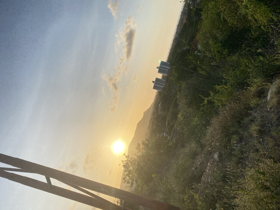 A sun-drenched landscape with buildings nestled into the side of a tree-covered mountain. The sun shines brightly behind the mountain, casting a golden glow over the sky and scenery. The lower portion of a wooden structure appears in the bottom left corner. The sky transitions from a pale blue to a yellowish hue. Wispy clouds are visible in the sky. The scene appears to be captured from an elevated vantage point.