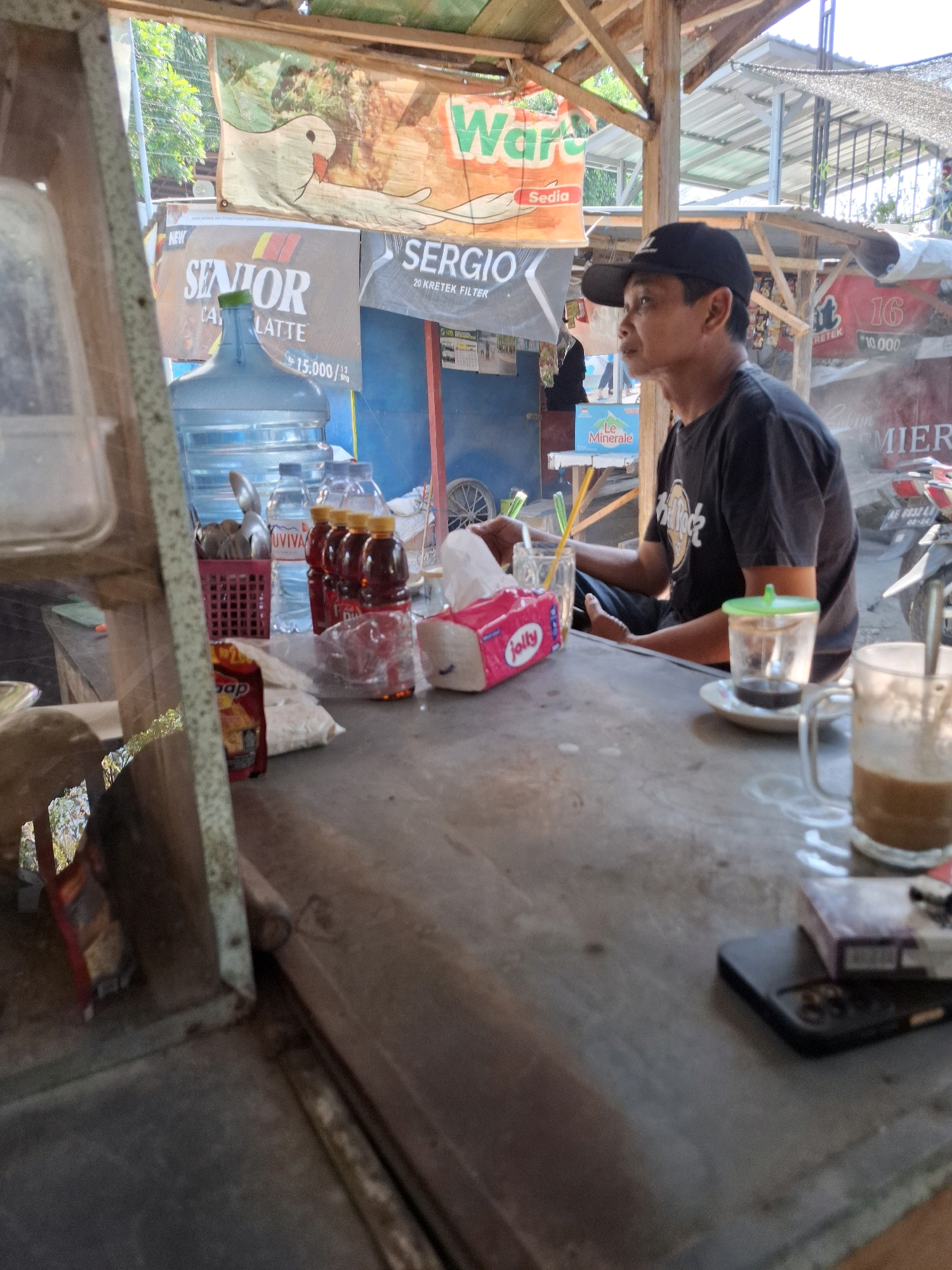 The image depicts an outdoor scene at a roadside stall in Tawun, Indonesia. A man wearing a black t-shirt and a black baseball cap is seated at a weathered table, looking to his right. On the table, there are several items including glasses of drinks, a pack of cigarettes, a mobile phone, a box of tissues, and several bottles of beverages. In the background, there are signs for "Warung Sedia," "Senior Cappuccino Latte," and other advertisements. A motorcycle is partially visible in the far right background. The stall appears to be constructed with wooden beams and a corrugated metal roof, suggesting a simple, open-air setup. The overall impression is that of a casual, informal marketplace or food stall.