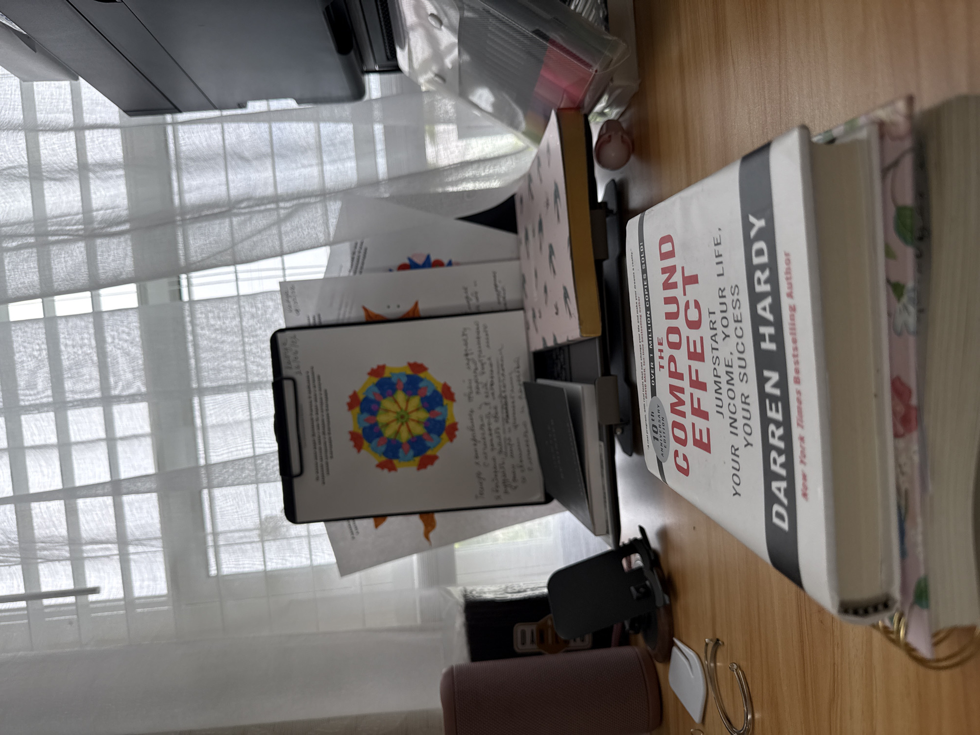 This is a close-up, eye-level shot of a wooden desk with several objects. The main subject is a book titled "The Compound Effect" by Darren Hardy. It is a thick, hardcover book with a white cover and red and black lettering. To the left of the book, there is a smartphone stand holding a smartphone. Behind the book and stand, there are several papers, some with drawings, clipped to a black clipboard. Further back, on the right side of the image, there is a stack of plastic-wrapped items, possibly markers or pens. A cylindrical speaker is visible on the far left of the desk, and a small pink object is in the background on the right. The desk is situated in front of a window with white curtains. The overall lighting suggests it is daytime. There are no people or animals visible in the image, and no specific location in Stock Island, United States of America, is identifiable from the scene.