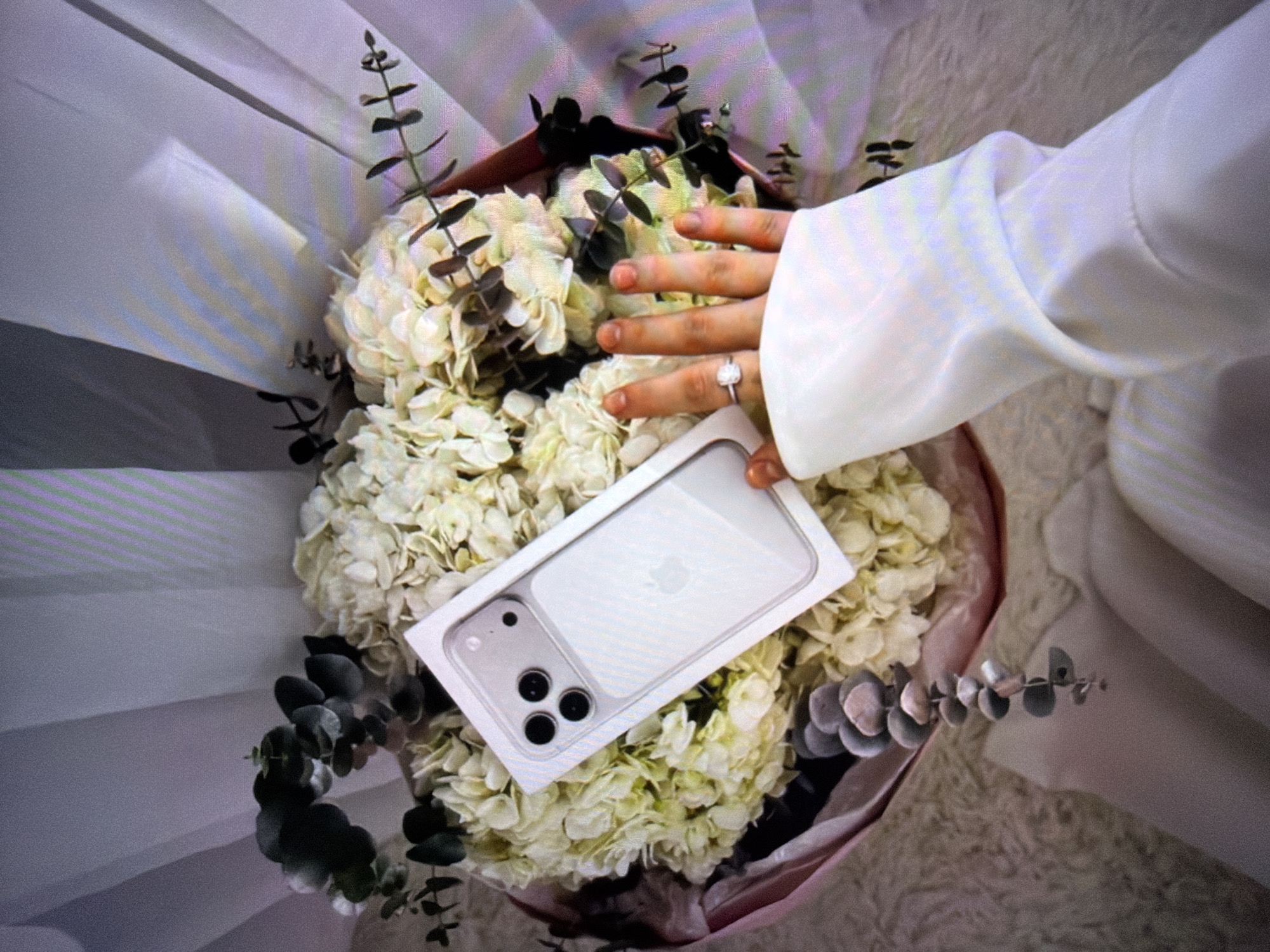 In Beșalma, Moldova, a bouquet of white hydrangeas and eucalyptus is presented. A person's hand, adorned with a square-cut diamond ring, rests on top of the bouquet, holding a white iPhone 14 Pro Max box. The background is out of focus, showing draped white fabric with vertical lines. The scene is captured from a top-down perspective.