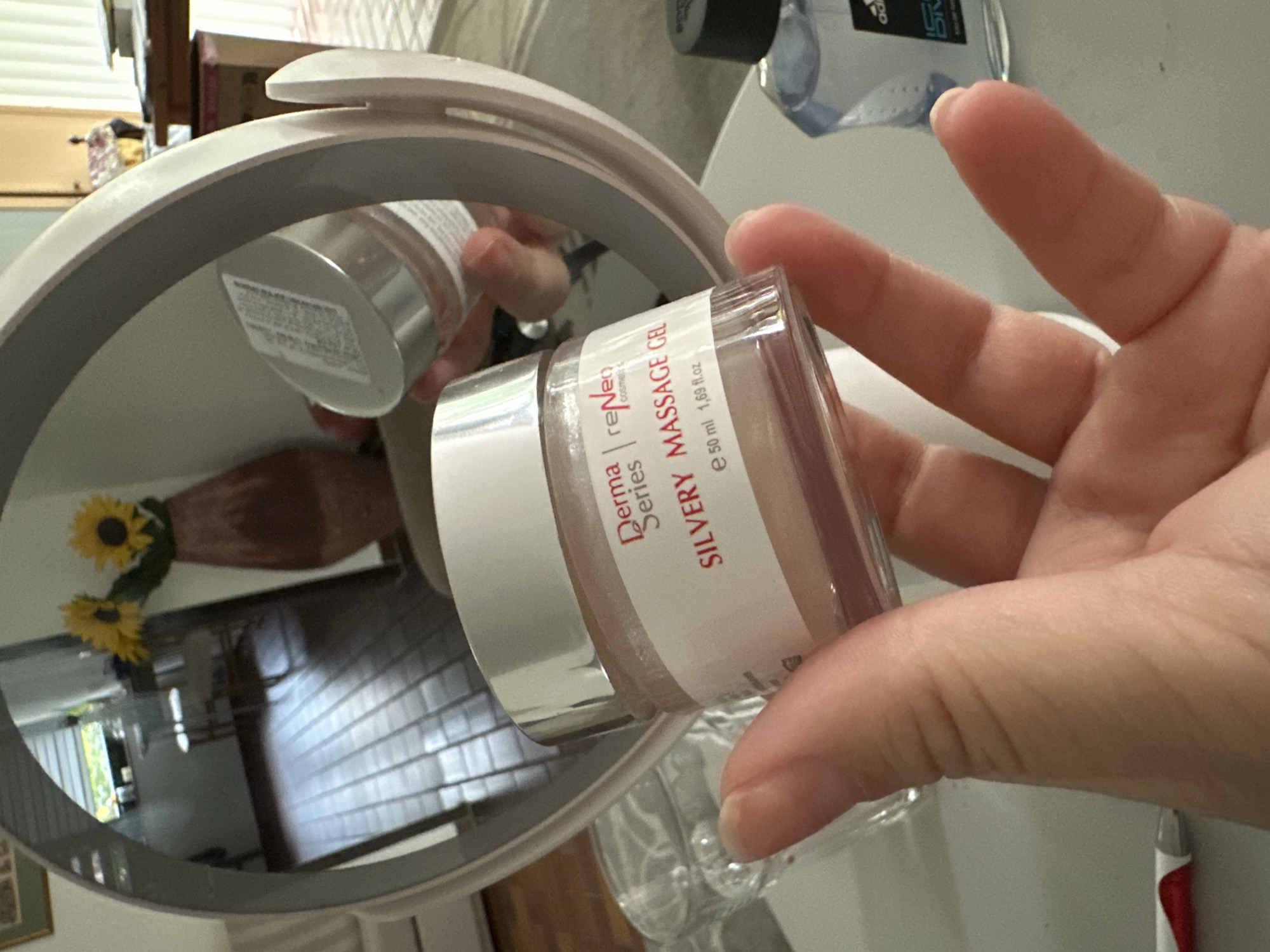 A close-up, eye-level, indoor shot shows a hand holding a clear glass jar with a silver lid. The jar contains a pinkish gel and has a white label that reads "Derma Series | reNeo COSMETICS SILVERY MASSAGE GEL e 50 ml 1,69 fl.oz". The background is a mirror reflection of a room with wooden floors, a window, and sunflowers in a vase. To the right of the jar, partially visible, is a clear blue bottle with a black cap, and to the left, a clear glass with liquid. The scene is likely in Krefeld, Germany, based on the context provided.