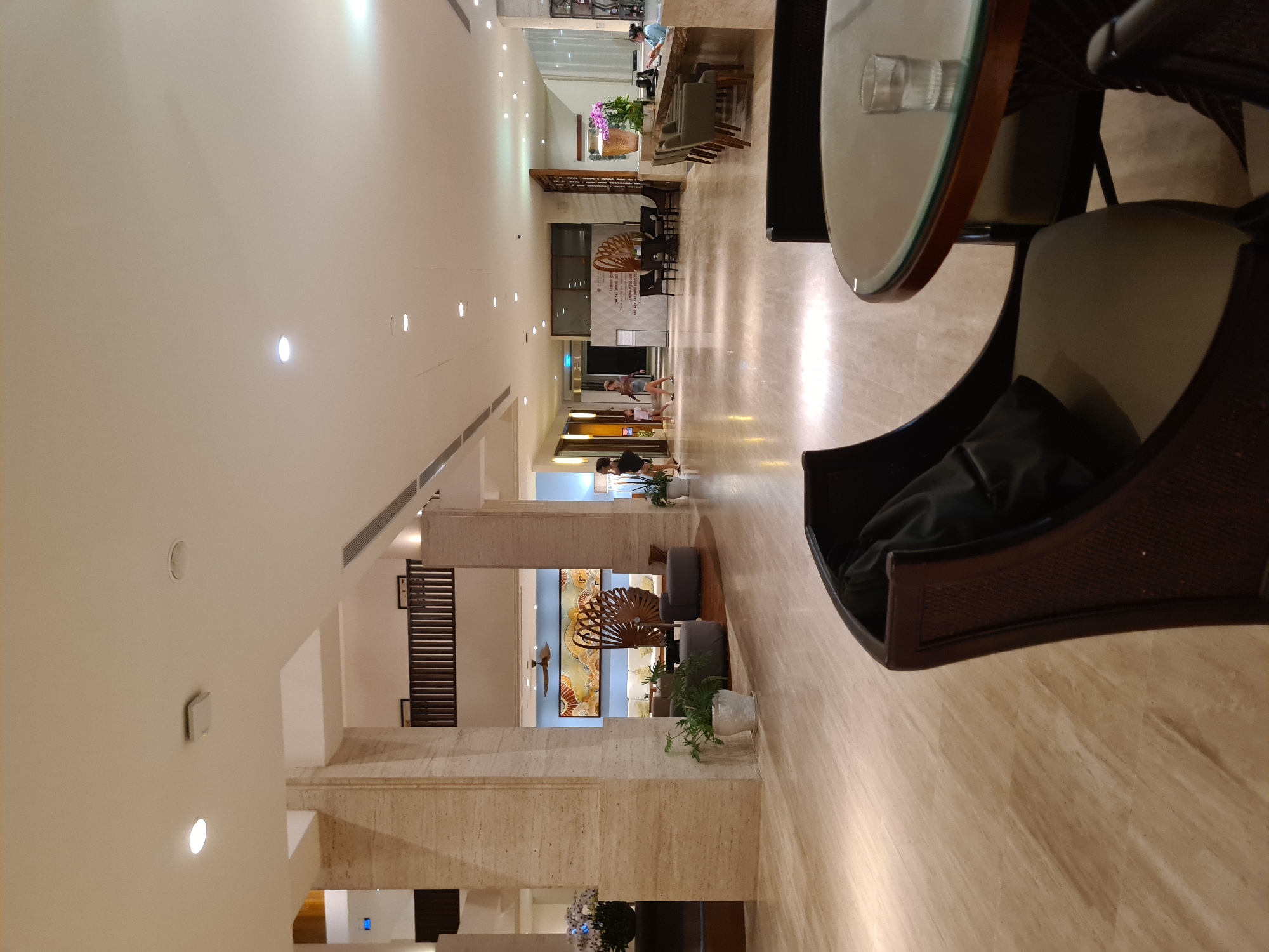 The image depicts the lobby of a hotel in Da Nang, Vietnam. A wide shot shows the polished marble floor reflecting the ambient lighting from the ceiling. In the foreground, a dark brown armchair with a black cushion is partially visible on the right side, and another similar armchair is on the left, facing a round glass-topped table with a single glass of water.
In the mid-ground, a reception desk is manned by a woman in a blue uniform, interacting with an unseen guest. Several high-backed chairs are arranged around the desk. Further back, a long, low reception counter is visible. To the left, a wall features a large abstract painting with shades of yellow, orange, and blue, and a ceiling fan. Two women and a child walk through the lobby towards the left of the frame.
The architecture features large, beige stone-clad pillars and a high ceiling with recessed lighting and air vents. The overall impression is one of a well-lit, spacious, and modern hotel lobby.