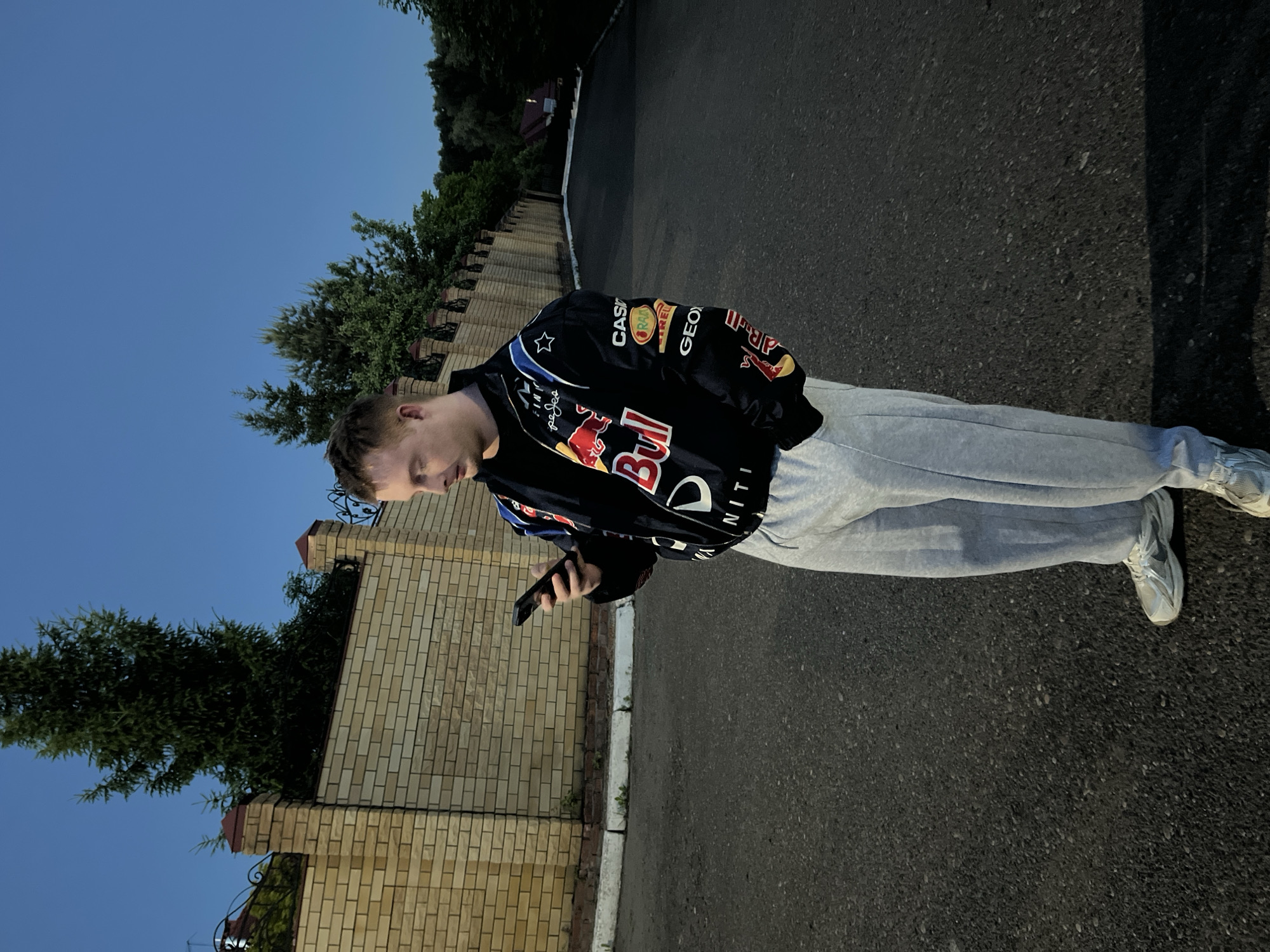 A young man stands outdoors, looking intently at a black smartphone held in his left hand. He is wearing a black racing-style jacket adorned with numerous colorful sponsor logos, including prominently visible "Red Bull," "CASIO," "Pirelli," "GEOS," and "INFINITI." He also wears light grey sweatpants and light-colored sneakers.
The setting appears to be a paved path or road, likely asphalt, with a white curb running alongside it. To his left, there is a tall brick fence or wall with dark coping and some visible ironwork. Behind the fence and extending upwards, lush green trees and bushes are visible. The sky above is a clear, bright blue, suggesting good weather.
The lighting indicates it's likely early evening or late afternoon, as the sky is bright but shadows are forming and the overall light quality is soft. The man's face is mostly obscured as he looks down, but he appears focused on his phone. The location cues, such as the brick fence and trees, suggest a suburban or residential area. Given the location context of Ореховка, Russia, this is likely a street or pathway in such a setting.
