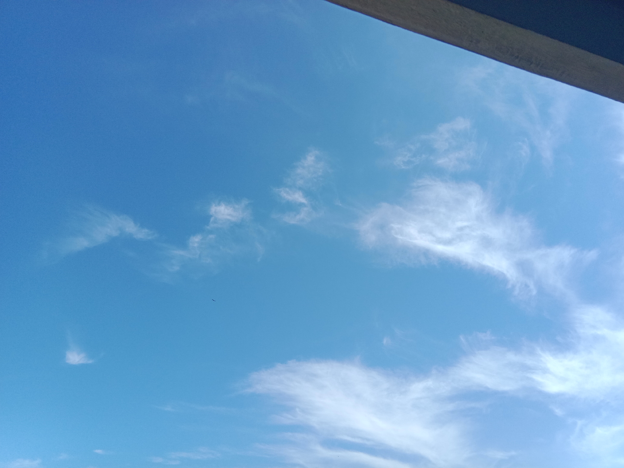 The image captures a bright blue sky with scattered wispy white clouds. A dark, angled object occupies the upper right portion of the frame, likely a roof or overhang, casting a slight shadow. In the lower left quadrant, a faint, small, dark silhouette of what appears to be a bird is visible against the sky. The image is devoid of any urban features of Leksono, Indonesia, presenting a natural outdoor scene.