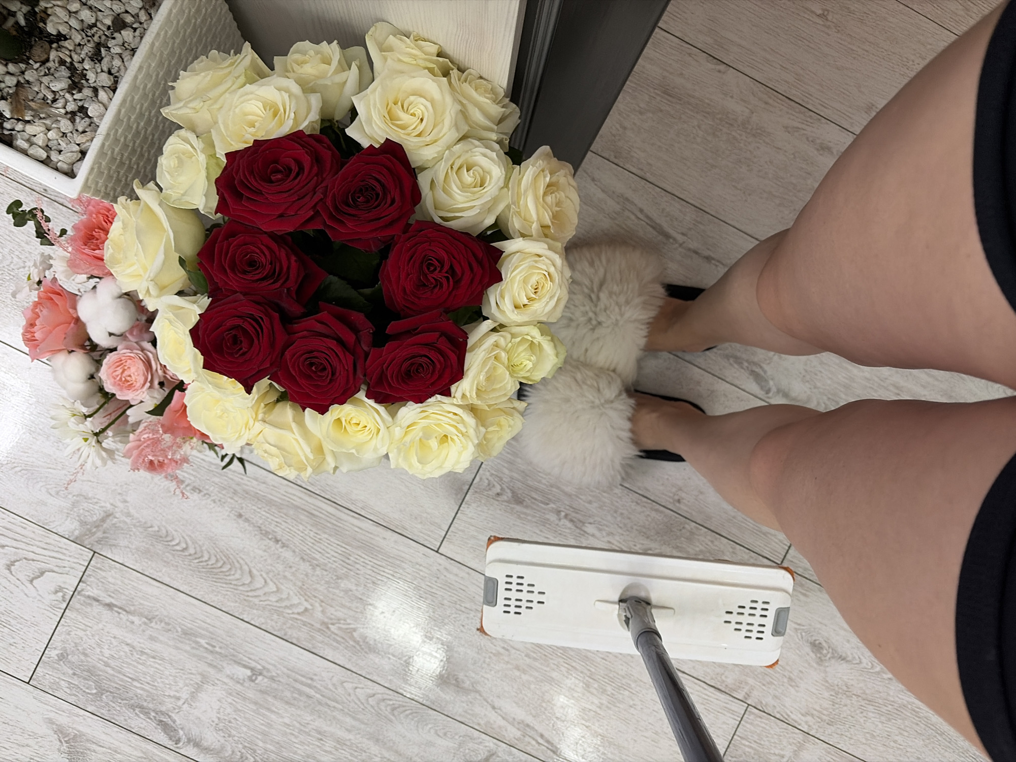 A high-angle shot shows a woman in slippers holding a mop, with a bouquet of red and white roses on a wood floor. The photo may have been taken indoors, possibly in a private residence. While the location in Chaikovsky, Russia, is mentioned, there's no visual information in the image to independently confirm this.