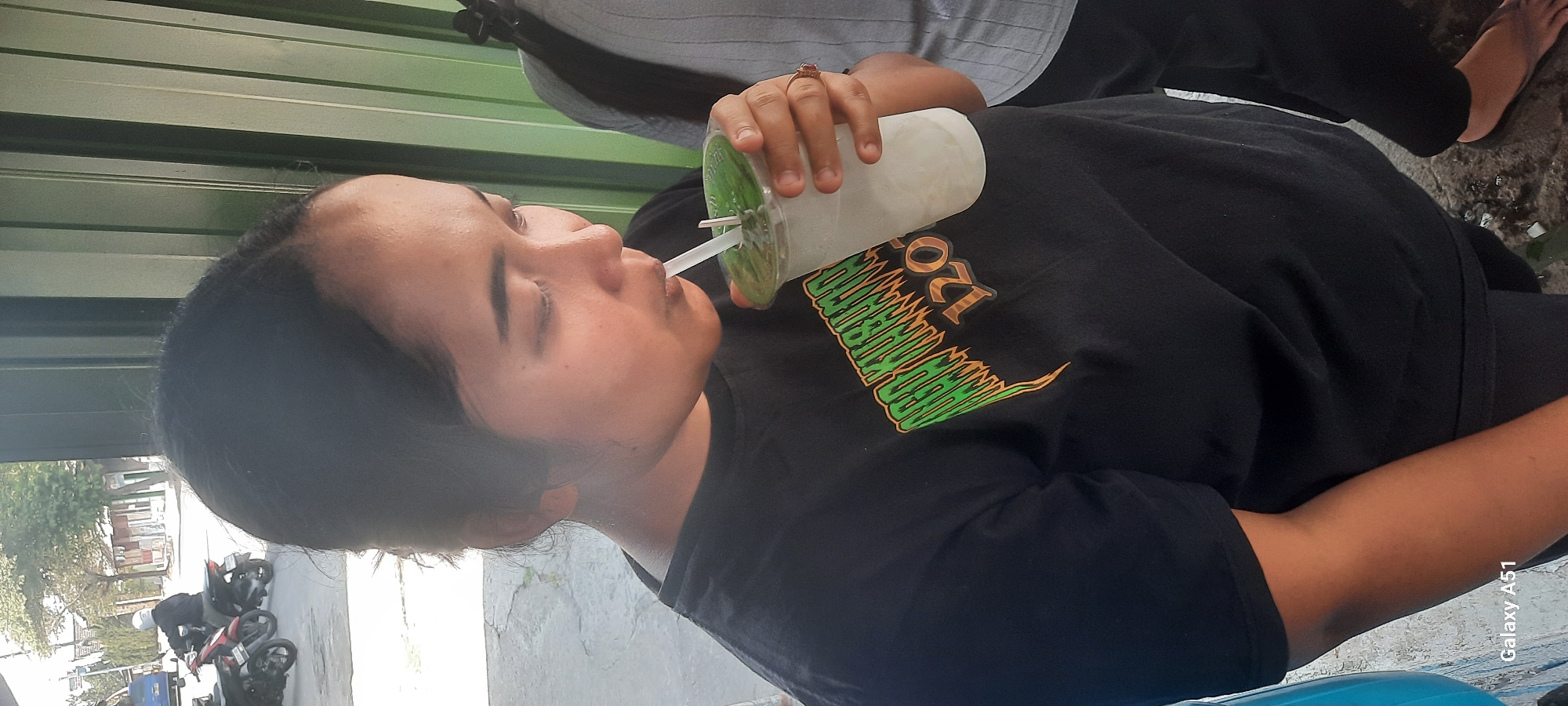 A young woman with dark hair, wearing a black t-shirt with a green and orange graphic design that reads "ARO KUSUMO 120", is captured in a medium shot outdoors, likely in Selopuro, Indonesia. She is holding a clear plastic cup filled with a white beverage and a straw, from which she is drinking. A silver ring is visible on her right ring finger. The background features a green corrugated metal wall. To the left of the woman, a blue object is partially visible, and further in the background, there is a street scene with motorcycles, including one ridden by a person wearing a white helmet. The ground appears to be a paved surface with some debris.