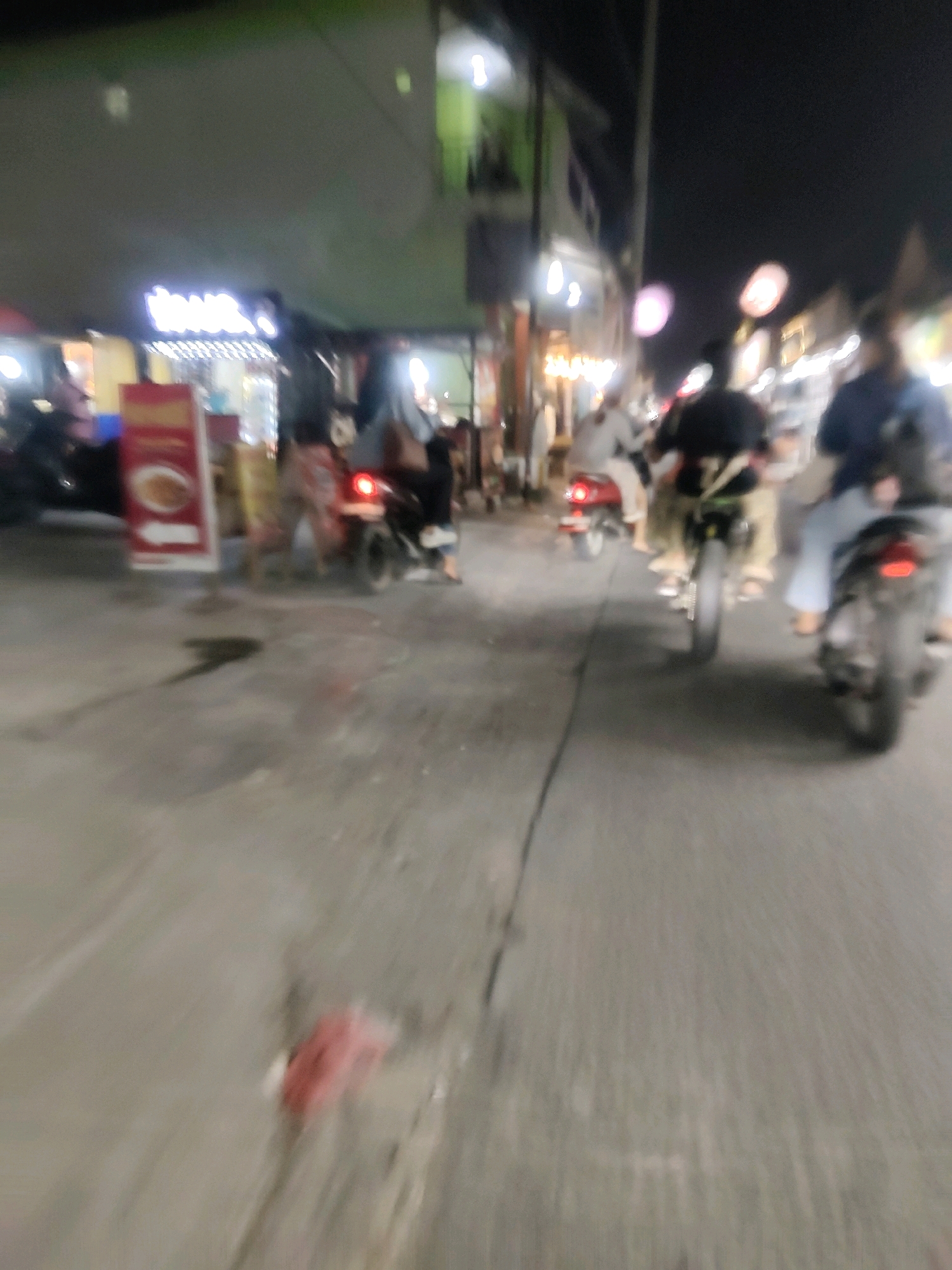 The image depicts a night scene on a street, likely in Mekarmukti, Indonesia, with motion blur indicating movement. Several people are riding motorcycles. On the left side, there are buildings with illuminated signs, one of which appears to be for a food establishment with a menu board displaying a bowl of food. The street is paved and appears wet in some areas. Some of the motorcycles have passengers. In the foreground, there is a dark, amorphous shadow on the ground and a reddish object. In the background, more buildings and streetlights are visible. The overall atmosphere is that of a busy street at night with local commerce and traffic.