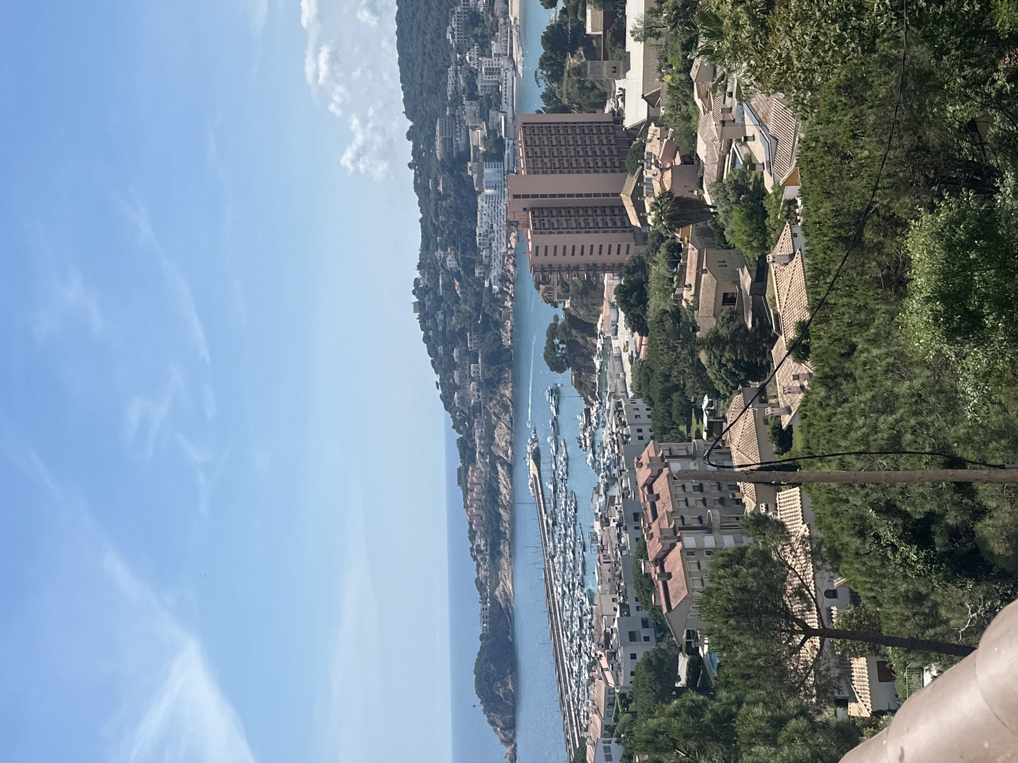 The image depicts a panoramic view of Sant Feliu de Guíxols, Spain, showcasing the coastal landscape and harbor. The sky is pale blue with scattered clouds. The sea is a deeper blue, with a long pier extending into the water, lined with numerous boats and yachts docked along both sides. The coastline is characterized by a rocky headland covered with vegetation and scattered buildings. The terrain slopes steeply towards the sea, with buildings and residences situated on the hillsides. The architecture is a mix of residential buildings and taller, possibly hotel structures, with terracotta roofs and light-colored walls. Green trees and vegetation are interspersed among the buildings, adding to the coastal landscape. In the foreground, a green area with trees frames the view.