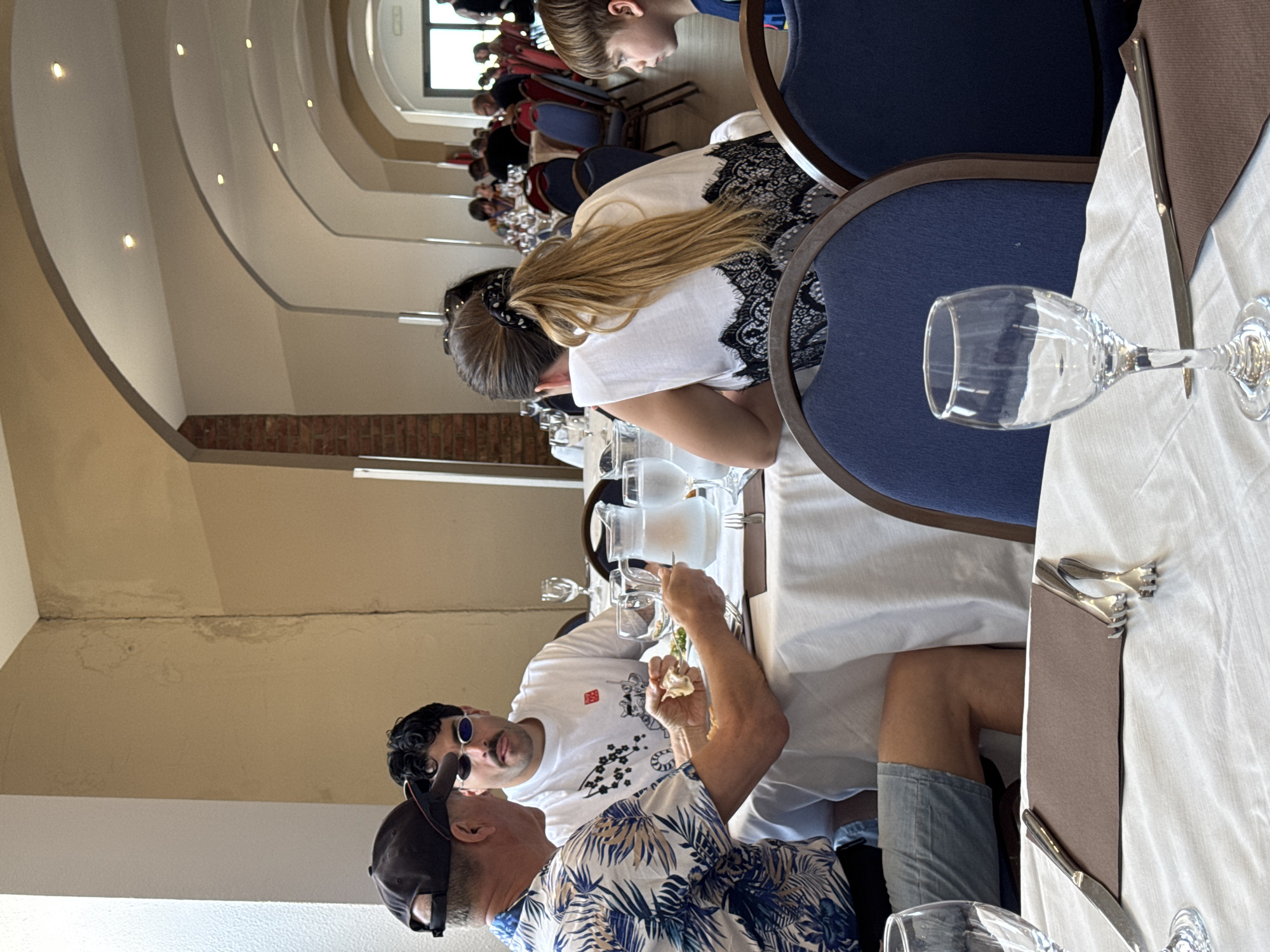 This image captures an indoor dining scene, likely in a restaurant or event hall in Isola delle Femmine, Italy. The setting features multiple tables covered with white tablecloths, set with clear glasses, cutlery, and brown napkins, and surrounded by blue chairs with dark wooden frames.
In the foreground, two men are seated at a table. One man, viewed from behind, wears a blue and white tropical print (Hawaiian) shirt and a dark baseball cap. He is actively eating, holding a fork to his mouth. Facing him is another man with dark curly hair and a mustache, wearing oval blue sunglasses and a white t-shirt adorned with black and red graphics, which include what appears to be Japanese characters and a stylized creature resembling a snake or dragon.
In the mid-ground, a woman with long blonde hair tied in a ponytail is visible from behind, wearing a white top with black lace detailing on the shoulders and back. Beside her, a young boy's head is partially visible. The background shows numerous other individuals seated at tables, suggesting a larger gathering or a busy dining period.
The room's architecture features curved, light-colored ceilings with recessed spotlights, creating a warm ambiance. Some walls incorporate exposed brickwork. Natural light appears to enter the space from windows visible in the far background. Notably, there is visible staining or discoloration on some sections of the upper walls and ceiling, indicating possible water damage. The overall atmosphere suggests a casual daytime meal or event.