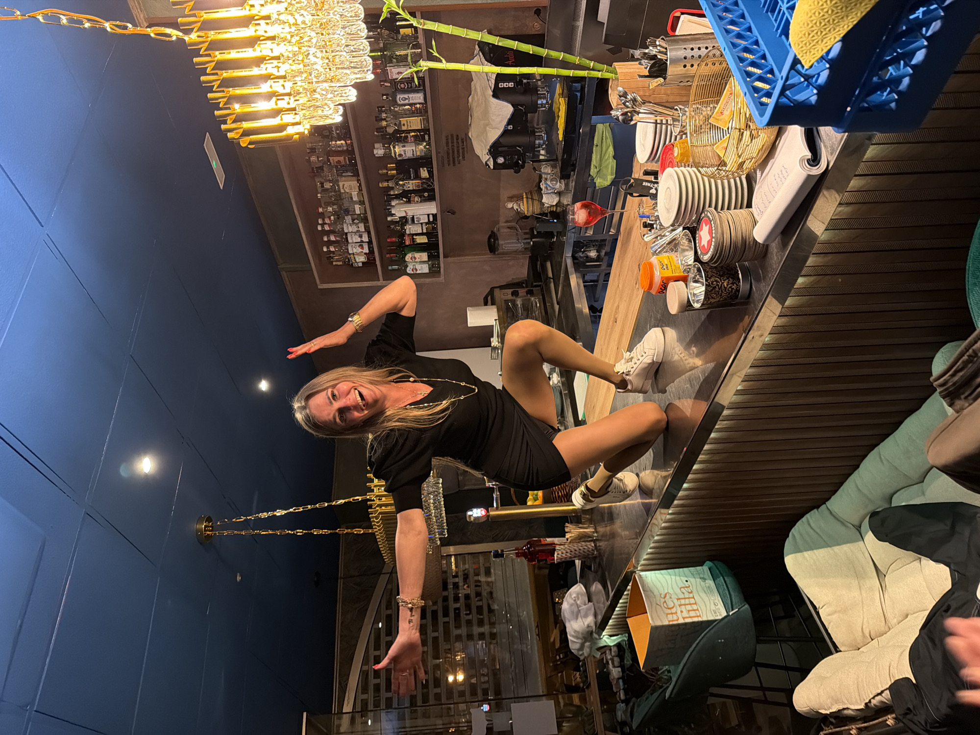 A woman is on top of a bar, likely in Salou, Spain. The bar is fully stocked with alcoholic beverages and bar equipment. She is wearing a short black dress and white sneakers. She has both arms outstretched. There is seating visible beneath the bar. The bar has horizontal striped decor.