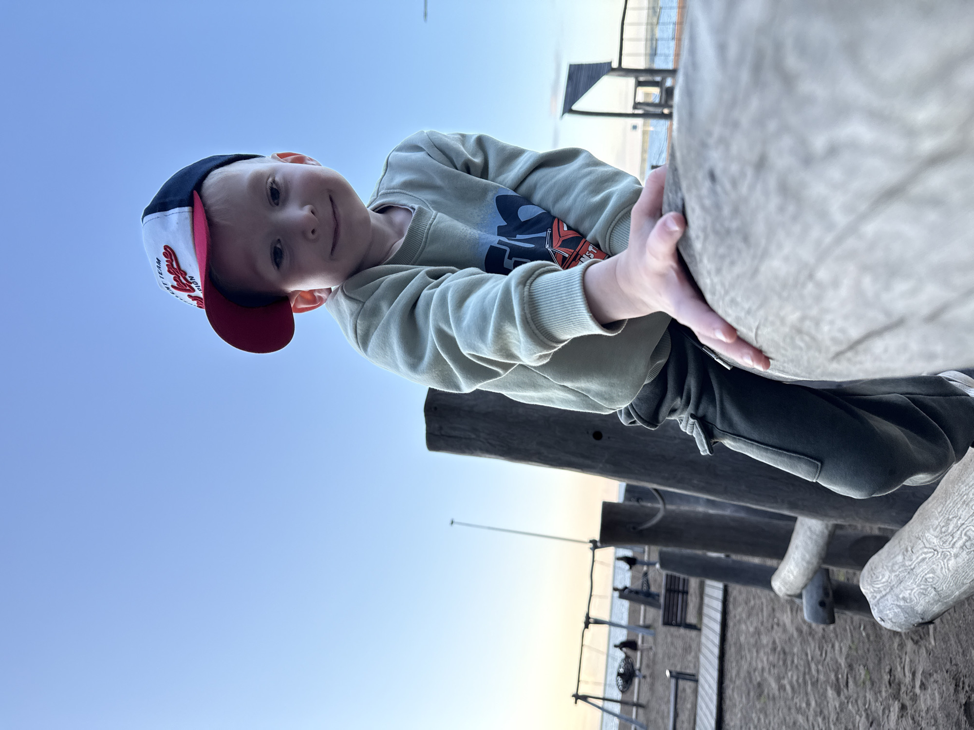 A low-angle photograph captures a young boy smiling directly at the camera, leaning over a large, textured, grey-brown wooden or concrete structure. His left hand grips the object firmly, with his right hand also resting on it. The image is taken from below, looking up towards the child and the expansive, clear blue sky above.
The boy wears a baseball cap with a white front panel, a red brim, and navy blue sides. Red embroidered text, partially visible on the cap, reads "TEAM CHAMPION." He is dressed in a light olive-green long-sleeved sweatshirt featuring a graphic design of an animated character or robot in black and orange on the chest. Dark sweatpants complete his outfit.
In the background, behind the child and the structure he is interacting with, other similar wooden or concrete elements suggest a playground or recreational area. Beyond these, a calm body of water (likely a lake or river) extends to the horizon, where faint structures, possibly part of a pier or boardwalk, are visible. The ground below appears to be sandy or dusty, indicating a waterfront setting.
The sky is a bright, clear blue, subtly lightening towards the horizon, characteristic of late afternoon or early evening. The lighting is soft and even. The child's expression is happy and playful, with a gentle smile and direct eye contact. This serene and cheerful scene is located in Chaikovsky, Russia.
