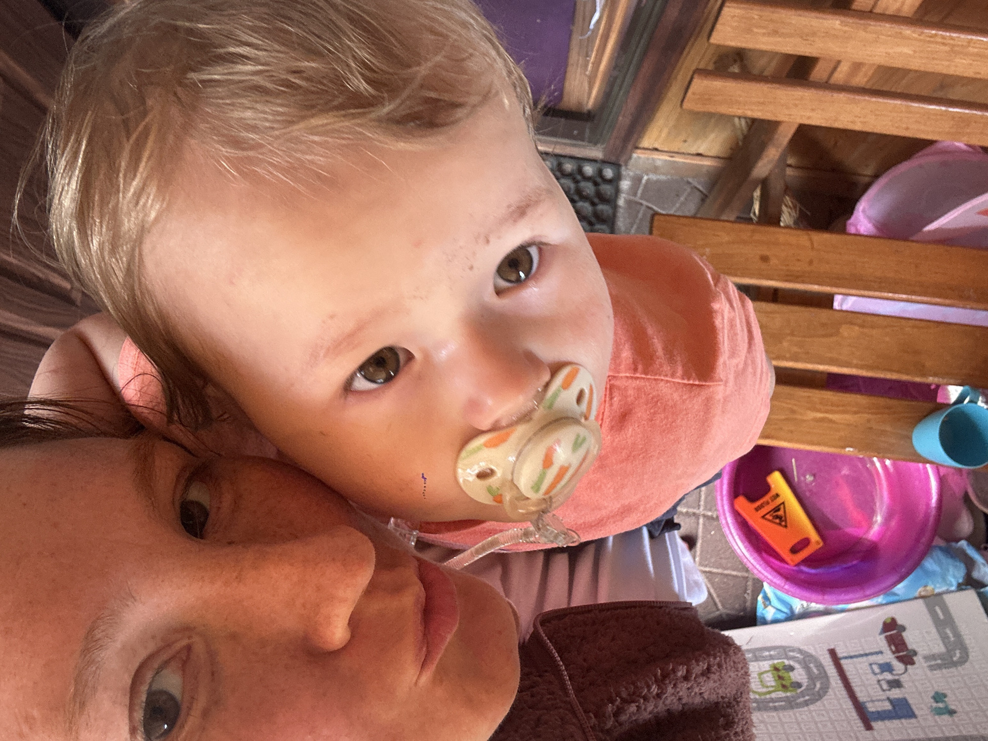 An eye-level, close-up shot depicts a woman and a baby. The woman has fair skin and gray-green eyes. The baby, who is sucking on a pacifier, is positioned above her with light brown eyes and fair skin. The baby wears a peach-colored shirt. In the background, a small yellow "wet floor" sign sits in a pink bowl next to a light-colored playmat. The location is not discernible from this image. The image is angled slightly upward.