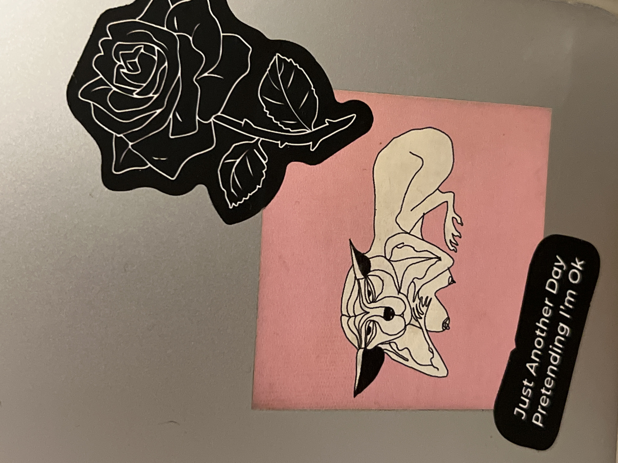 The image displays a close-up view of a silver-grey surface, likely a laptop lid or similar object, adorned with three distinct stickers.
In the upper left, a large, black sticker shaped like a rose is visible. It features intricate white outlines detailing the petals of a blooming rose and two leaves on a stem, against the solid black background of the sticker.
To the right and slightly below the rose sticker, there is a rectangular pink sticker. This sticker features a stylized line art drawing of a figure. The figure is depicted with a cream-colored body outlined in black, appearing to be in a crouched or reclined position. It has distinct features resembling pointed ears or horns, a humanoid torso with visible nipples, and abstract limbs. The drawing occupies most of the pink sticker's surface.
In the bottom right corner, a small, black, rounded rectangular sticker is oriented vertically. White text is clearly legible on this sticker, reading: "Just Another Day Pretending I'm Ok".
The overall lighting is even, suggesting an indoor setting without harsh shadows. The silver-grey background surface shows a slight texture. No specific activity is taking place, as the image is a static shot of the decorated surface.