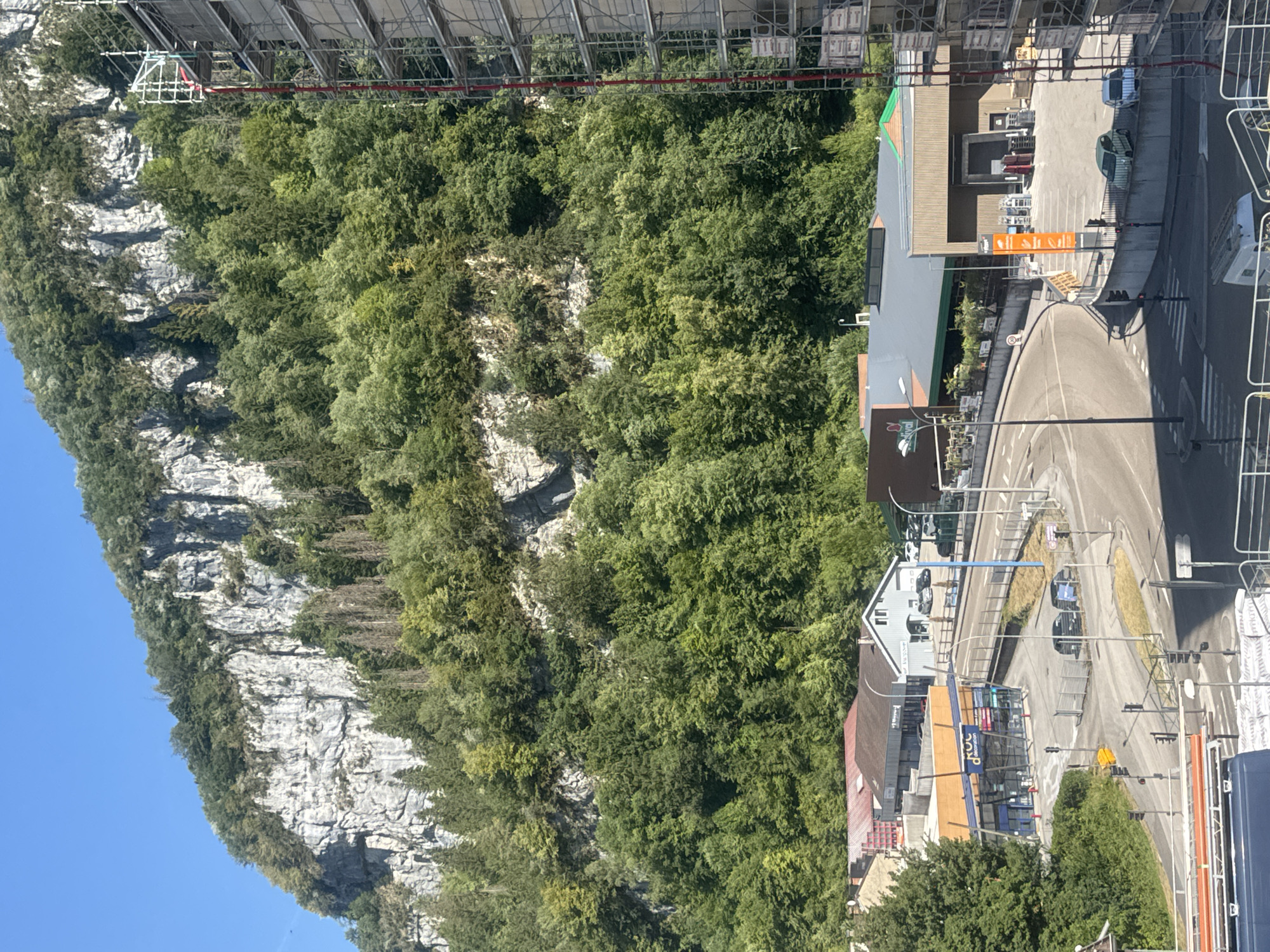 The image depicts an aerial view of a commercial area in Saint-Claude, France, nestled at the base of a steep, tree-covered cliff face. The cliff is a prominent natural feature, its rocky surface exposed in places and heavily forested on its slopes. In the foreground, a modern street curves through the town, with cars visible on the road and in parking lots. Several commercial buildings are present, including what appears to be a store with an orange facade and blue signage, and a larger building with a grey roof and a loading bay. Scaffolding is erected on the side of a multi-story building on the right side of the image, suggesting ongoing construction or renovation. The sky is clear and blue, indicating a sunny day. The overall scene conveys a sense of a small town integrated with its natural surroundings.