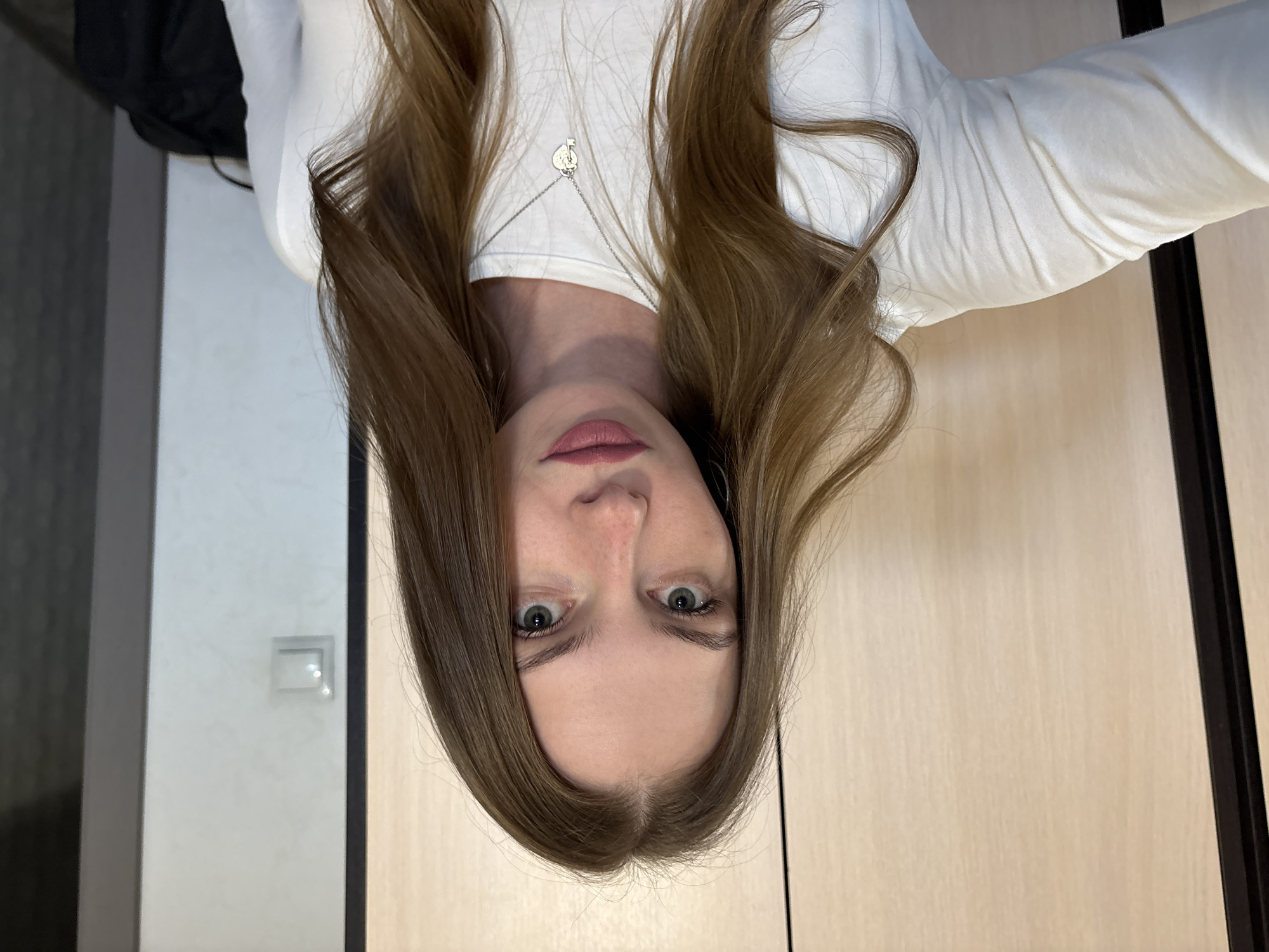 A young woman with long, light brown hair and green eyes is shown in a close-up, self-taken photograph. She is wearing a white, long-sleeved shirt and a silver necklace with a heart and key pendant. Her lips are painted a matte, dusky pink. The background consists of a light wood-paneled wall on the left and a light gray wall with a light switch on the right. The lighting appears to be natural, coming from the front. The image is captured indoors, likely in a residential setting. The city of Perm, Russia is not visible in this image, nor are any elements that specifically indicate the location.