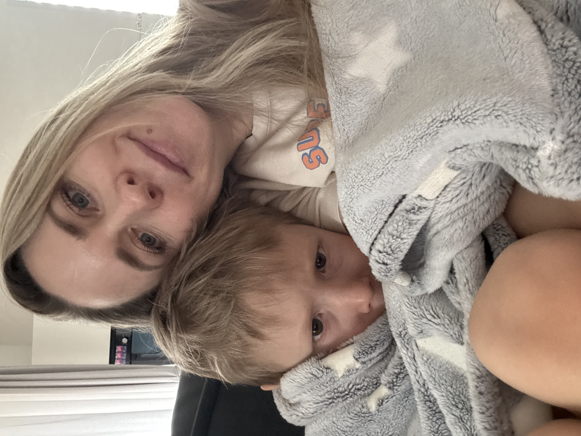 A woman with blonde hair and blue eyes is seen from an upside-down perspective, along with a young child covered in a gray blanket with white stars. The woman is wearing a light-colored sweatshirt with orange lettering. The background is indistinct, but a window and a computer screen can be partially seen. The location of the image is unknown as it is not mentioned in the prompt.