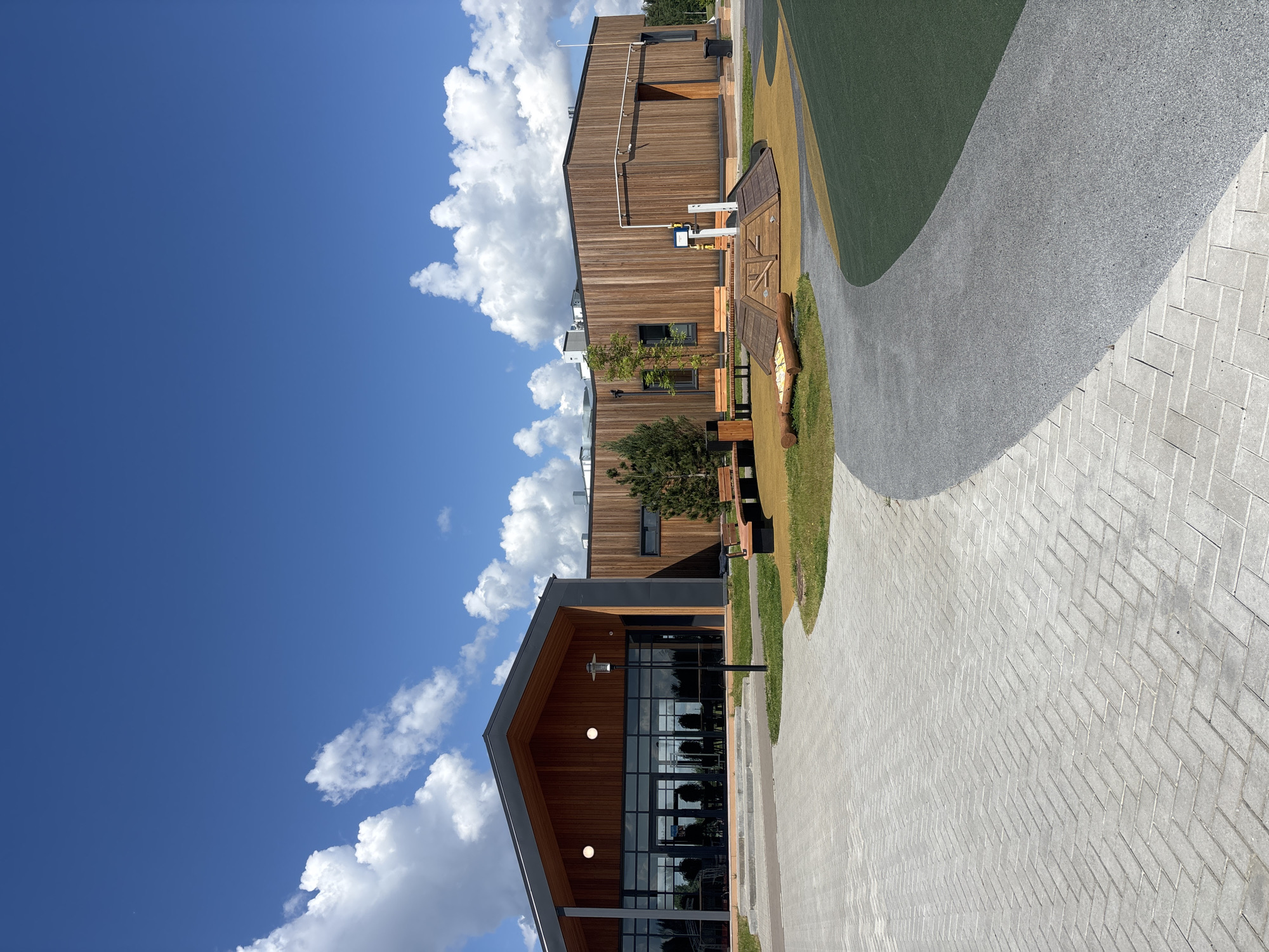 The image depicts a modern building complex located in Varaksino, Russia, under a bright blue sky dotted with puffy white clouds. The buildings are constructed with wooden siding and feature large windows. A paved walkway, laid in a herringbone pattern, leads up to one of the buildings. Adjacent to the walkway is a strip of green grass and a gray path. A wooden bench is also visible. The scene appears to be a well-maintained outdoor area, possibly a park or a courtyard within the building complex. There are no people visible in the image.