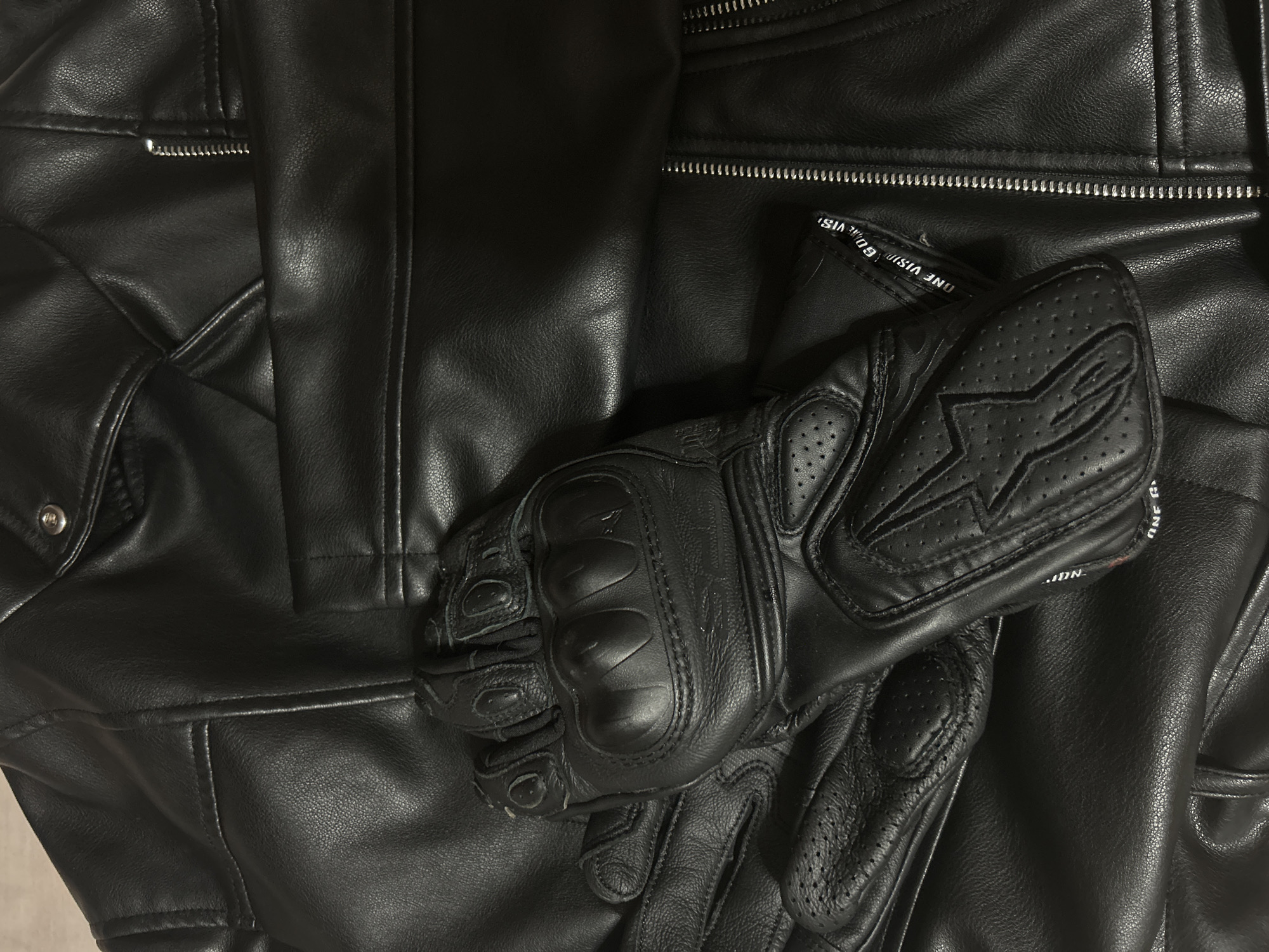 The image displays a pair of black leather motorcycle gloves placed on top of a black leather jacket. The gloves are the main subject and are positioned centrally. The leather jacket serves as a textured background. The location is not specified.