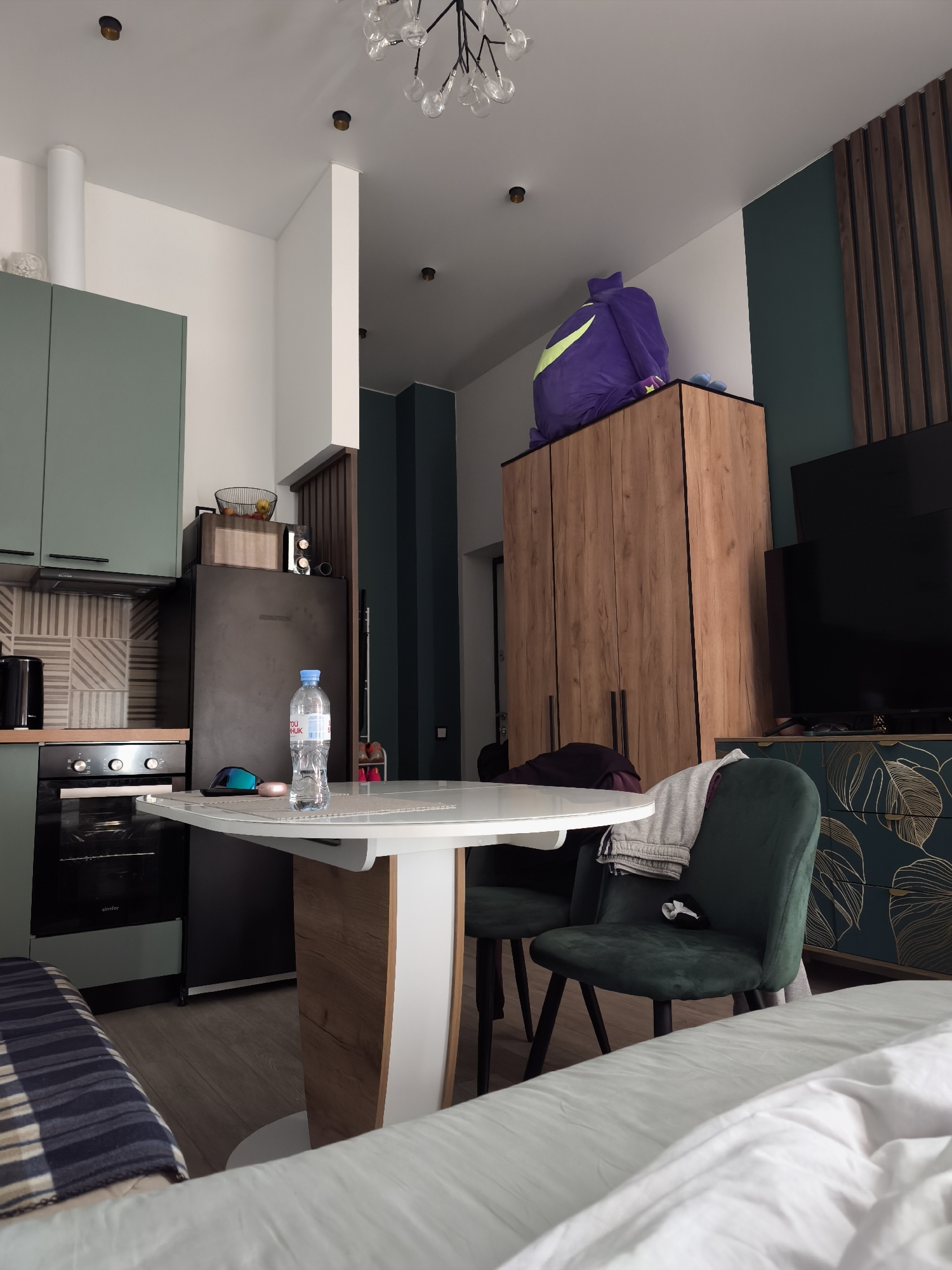 This interior shot depicts a room, possibly a studio apartment, with kitchen and living areas. The kitchen area features green cabinets and a dark-colored refrigerator, with a microwave and some decorative items on top of the fridge. The kitchen also contains an oven. A round white table with a wood-grain accent is positioned in the center of the room, with two green chairs.
The living area has a bed with white linens. The style of the furniture indicates that the location is likely to be in a modern apartment, and the architecture and furnishings are suggestive of Moscow, Russia.
A tall wooden wardrobe is located along one wall, with a large purple bag placed on top. Next to the wardrobe is a television placed on a cabinet. The television and cabinet are next to an adjacent dark green wall with wood slat accents. The room is lit by ceiling lights.