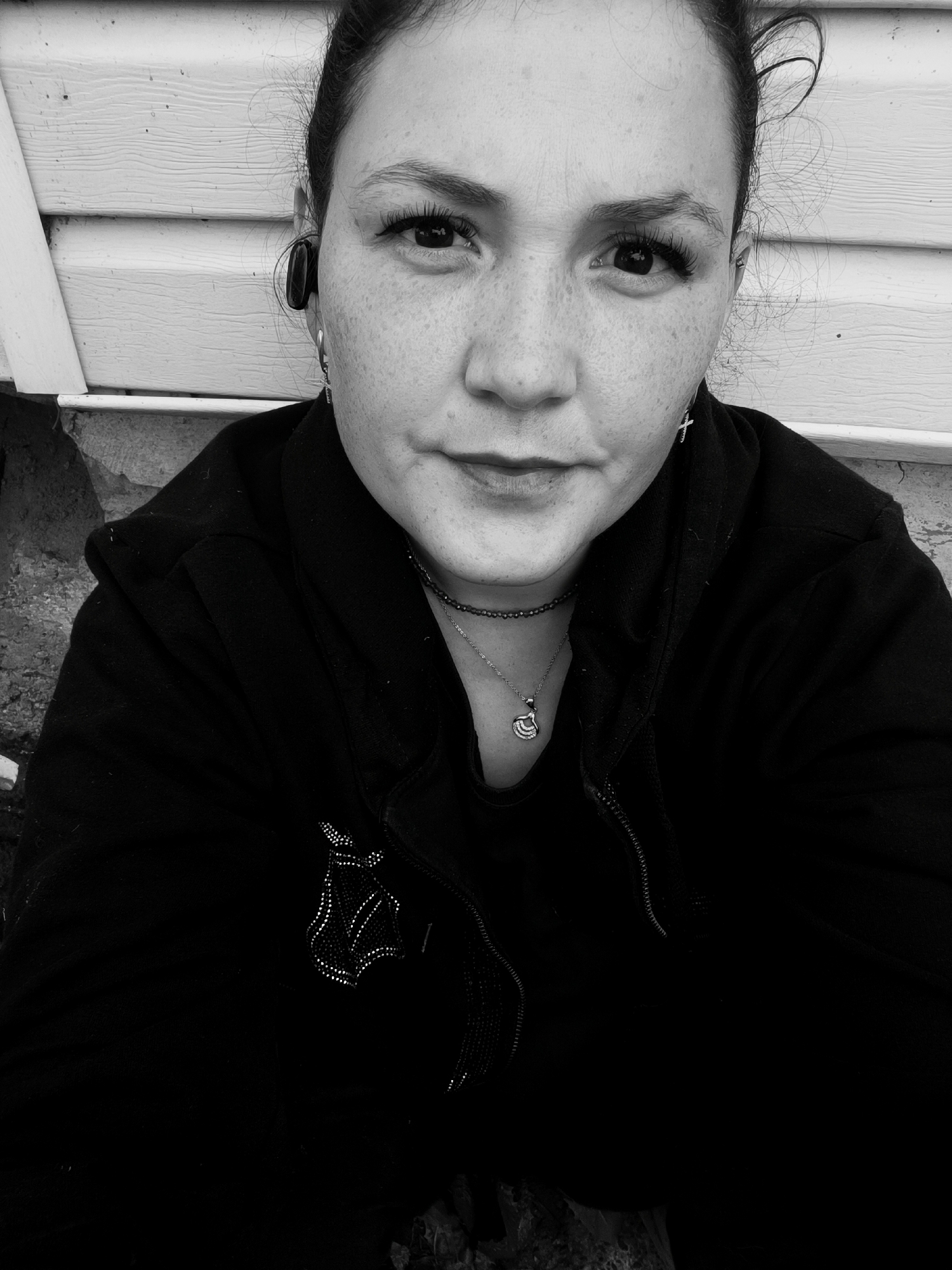 A black and white, waist-up, selfie-style photograph shows a woman with dark hair pulled back. She is wearing a black hooded jacket with a zipper and a silver pendant necklace. She also has a small silver cross earring in her visible ear. The background consists of a light-colored wooden wall and a dark, textured surface, possibly a stone wall or embankment. The image is not specifically located in the city of Karsha, Russia.