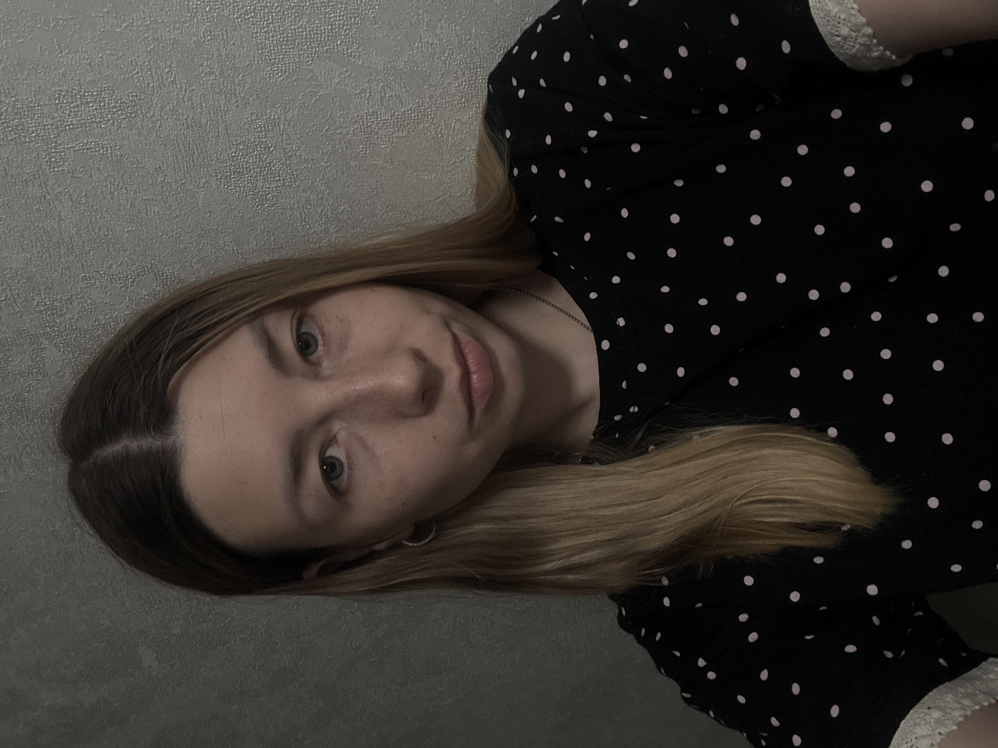 A woman with blonde hair is facing the camera. She has fair skin, blue eyes, and freckles. She is wearing a black dress with white polka dots. A small silver earring is visible in her left ear. The background appears to be a textured grey wall. No information in the photo indicates that it was taken in Moscow, Russia.
