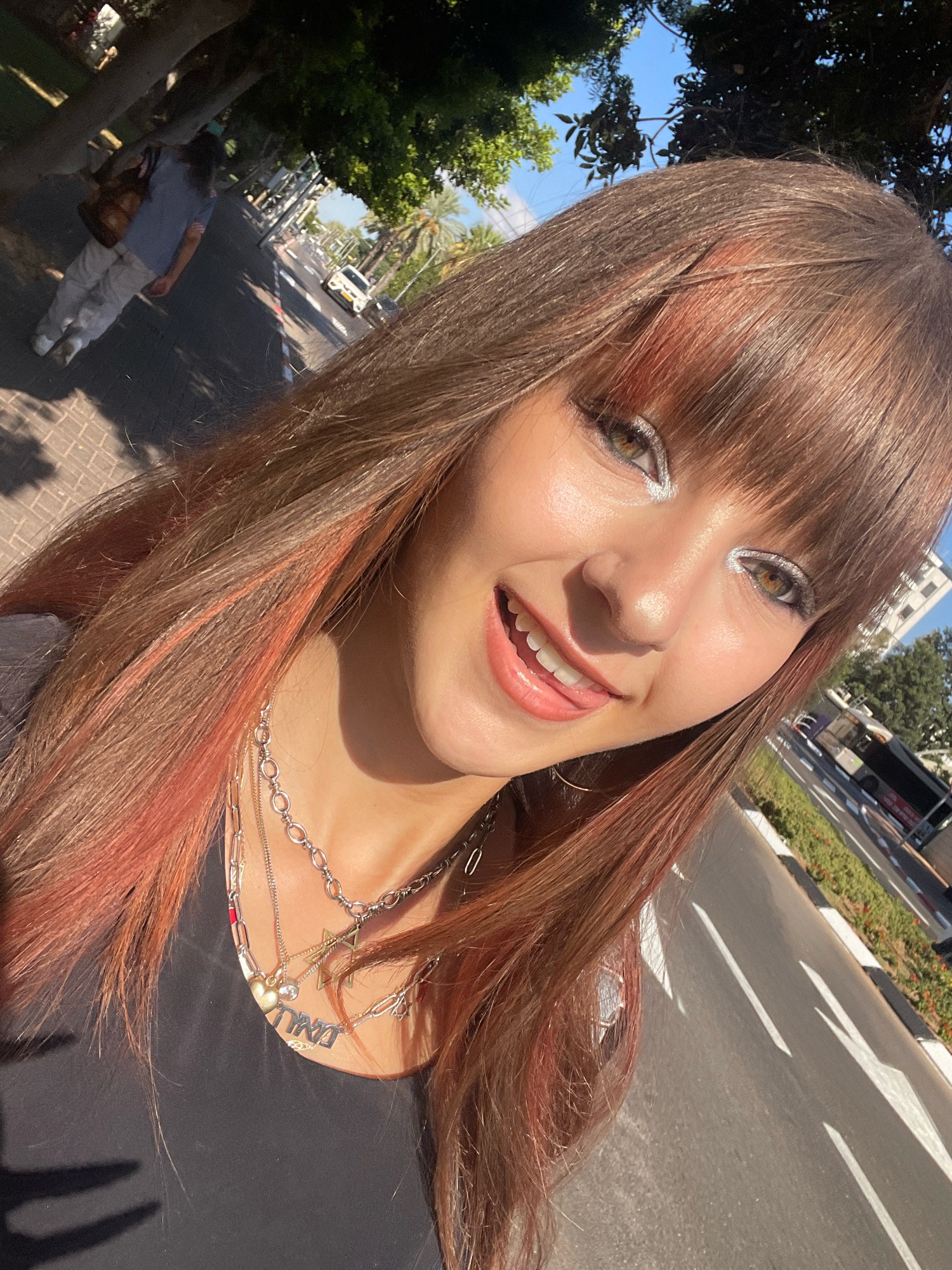 The image shows a young woman taking a selfie outdoors on a sunny day, with a street and sidewalk visible in the background. She has long, brown hair with streaks of pink, and is wearing silver eyeshadow and lip gloss. She is also wearing a black top and several necklaces, one of which has a pendant with Hebrew writing.
In the background, a person in a light-colored shirt and pants is walking away from the camera on a sidewalk. A street with cars and a bus is visible further down the road, lined with palm trees and buildings. The overall scene suggests a casual, urban environment in Raanana, Israel, likely during daylight hours given the bright sunlight and clear blue sky.