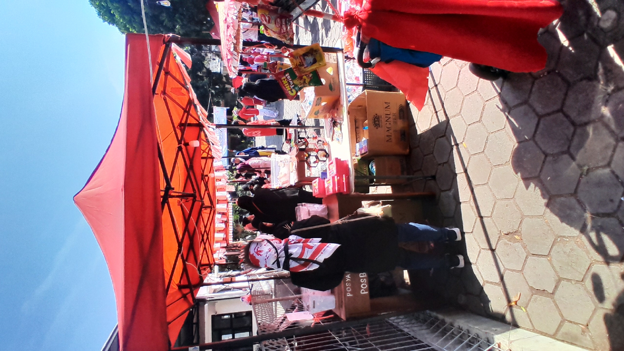 An outdoor market is depicted under a bright, clear sky with abundant sunlight. Red canopies are erected over stalls, casting shadows on the hexagonal patterned pavement below. A person wearing a black coat and jeans, with a white and red headscarf covering their face, stands facing away from the camera. They are positioned in front of a table displaying various items, including clear containers and pink boxes. Behind this individual, several other people are visible, some browsing at stalls laden with goods such as snacks, boxes, and bags. Further in the background, more stalls and market activity can be observed. A metal fence lines the left side of the scene. The overall impression is of a bustling, daytime market event.