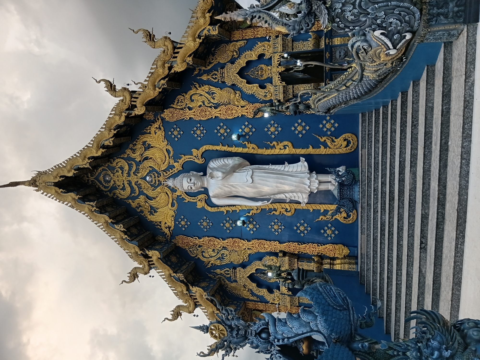 The image shows the Wat Rong Khun, also known as the White Temple, in Chiang Rai, Thailand. It is a contemporary, unconventional, and privately owned art exhibit in the style of a Buddhist temple. The temple is adorned with intricate blue and gold details and features a large white Buddha statue in front of the main building. The roof is decorated with mythical creatures and golden embellishments. The foreground shows a set of wide stone stairs leading up to the temple, flanked by large, dark blue sculptures of mythical creatures with golden accents. The sky above is overcast with a muted, diffused light.