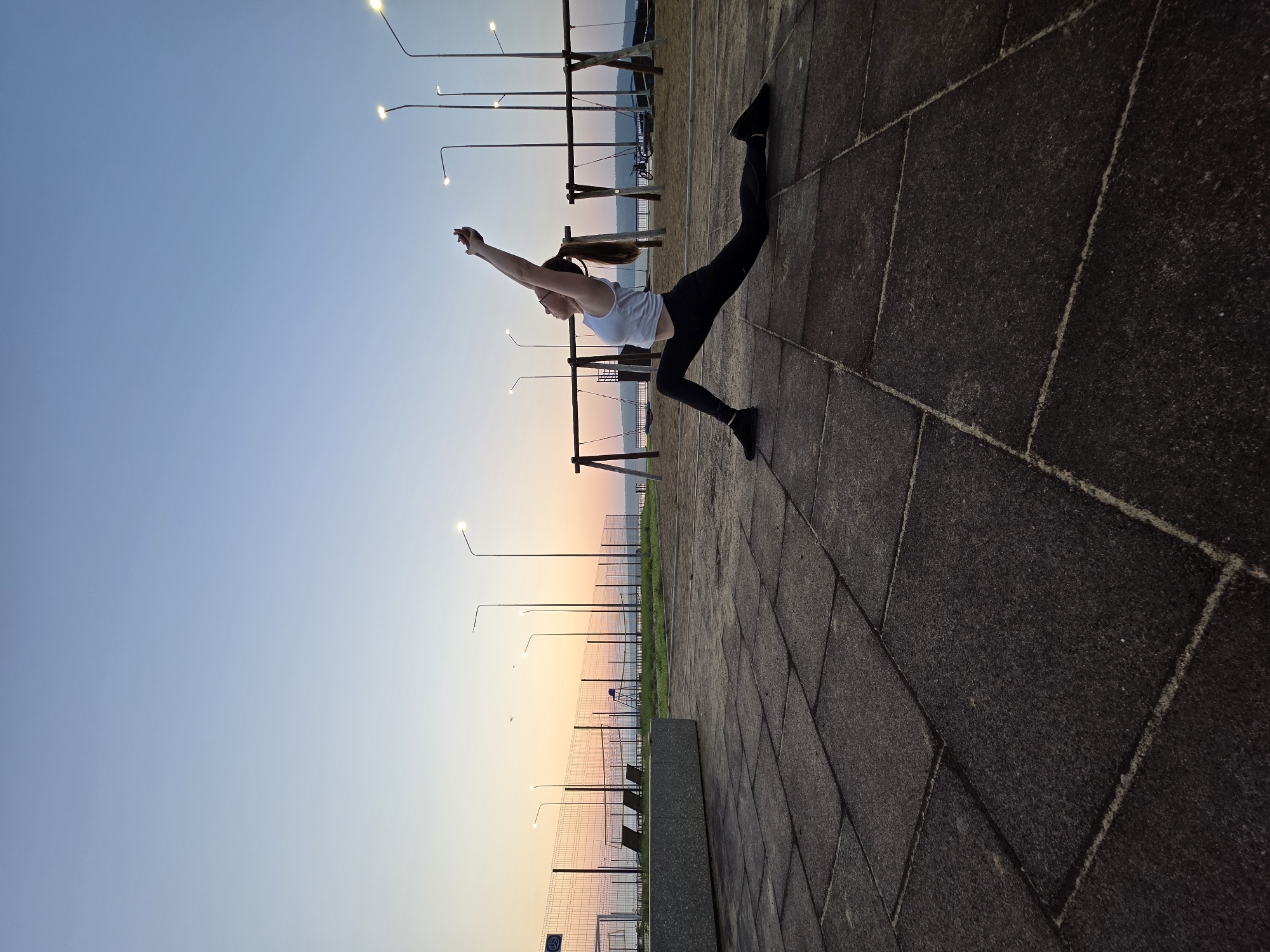 A woman is practicing yoga outdoors on a paved surface at dawn. She is in a low lunge pose with her arms raised and hands clasped overhead. The woman is wearing a white tank top and black leggings. The background shows a fence, swings, streetlights, and a body of water. The sky is a gradient of pale blue and orange. The location appears to be a public recreation area, possibly a park or playground, in Chaikovsky, Russia.