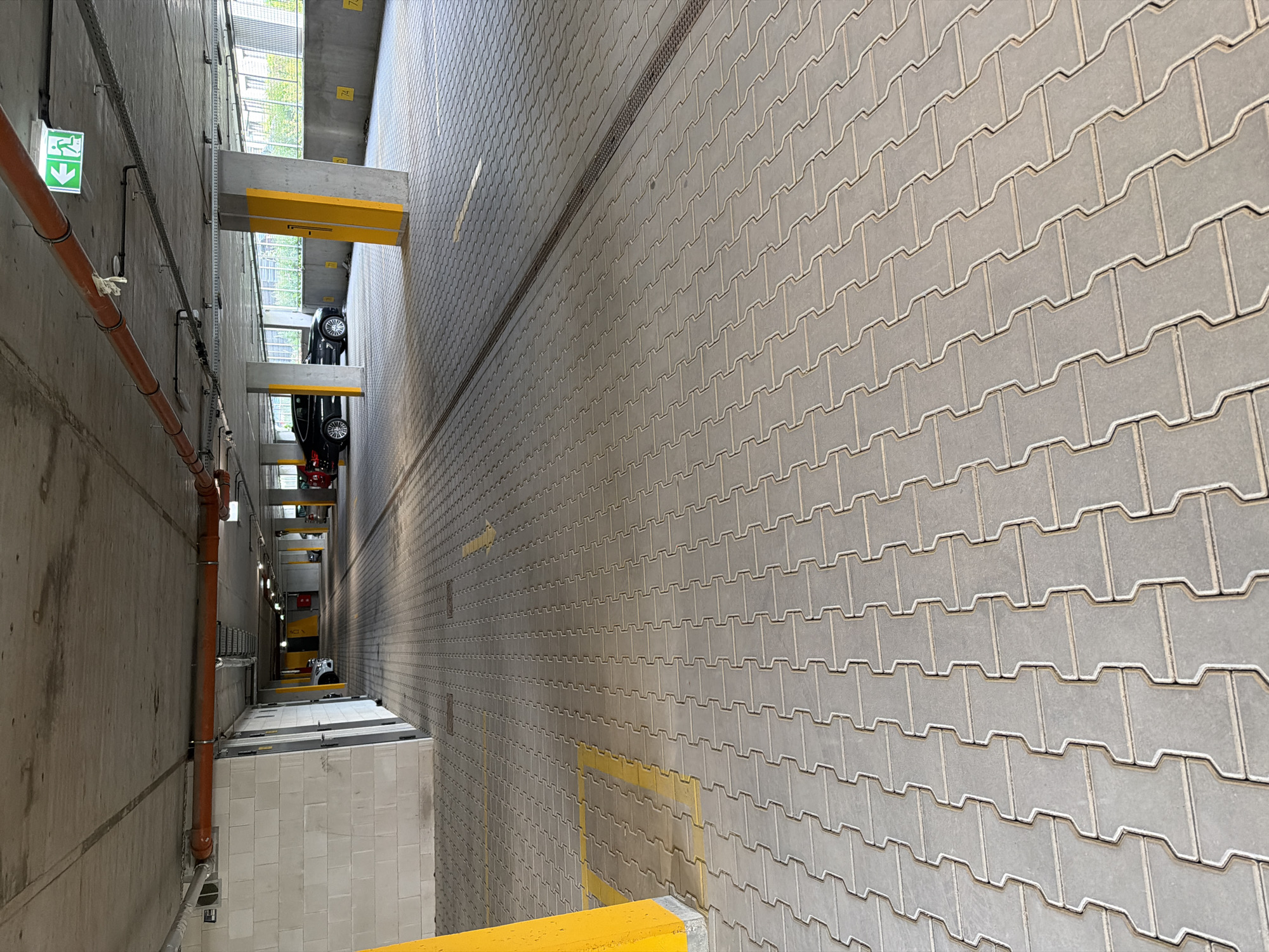 The image shows a section of an indoor parking garage. The floor is paved with interlocking grey concrete pavers. Yellow painted lines are visible on the floor, indicating parking spaces and directional arrows. Several concrete pillars, some painted yellow at their base, support the ceiling. Pipes and conduits are suspended from the concrete ceiling. In the background, a few cars are parked, and an emergency exit sign with a green arrow pointing left is illuminated on the ceiling. The overall environment is that of a modern, functional parking facility. The image does not provide any specific details to locate it within the city of Łódź, Poland.