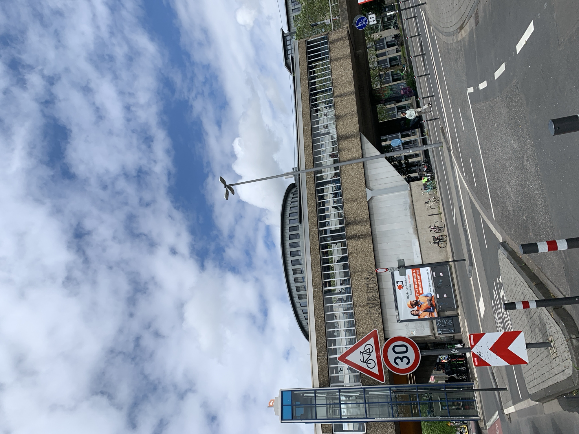 This image captures an urban street scene during the daytime, with the perspective rotated 90 degrees clockwise, placing the sky on the left and the street on the right when viewed upright. The sky is largely blue with scattered white, wispy clouds.
In the foreground and midground, a multi-story building dominates the left half of the image. It features a facade of concrete or stone with numerous rectangular windows, many reflecting the sky. Part of the building has a distinctive curved, possibly metallic, roofline. Below this structure, another section with glass walls and a blue-tinted frame is visible, possibly an entrance or part of a public transport stop.
Various street signs are prominent. A triangular warning sign with a red border and a bicycle symbol is visible, alongside a circular speed limit sign displaying "30". Further to the right, a red and white chevron sign indicates a curve to the left. On the road, a blue circular sign with a white bicycle and double arrows (↓↑) suggests a bi-directional cycling path.
The right side of the image features a paved road with white lane markings and a textured sidewalk area. Several black and red/white striped bollards line the edge of the road. Bicycles are parked near the building, and a few indistinct figures can be seen walking or through the building windows. A street light with a distinctive, multi-faceted lamp head stands tall near the building.
A large billboard is affixed to the building, displaying text in German for a personnel service. The text reads: "EDO Personalservice G", "Einfach Gut. Vermittelt", "JETZT BEWERBEN & DURCHSTARTEN!", and "edo-personalservice.de". The weather is bright and sunny with visible clouds.