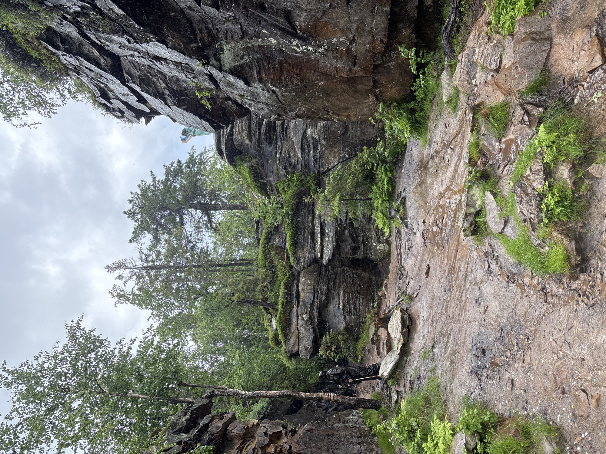 The image shows a rocky, forested area with a cloudy sky, suggesting it is raining. The terrain is uneven and muddy, with rocks and vegetation. In the foreground, two people are walking on a path, wearing rain gear and carrying hiking poles. In the background, on a higher rock formation, a person is standing and looking at a painting or artwork. The scene appears to be a natural landscape, possibly a hiking trail or a park, located in the city of Nagornskiy, Russia.