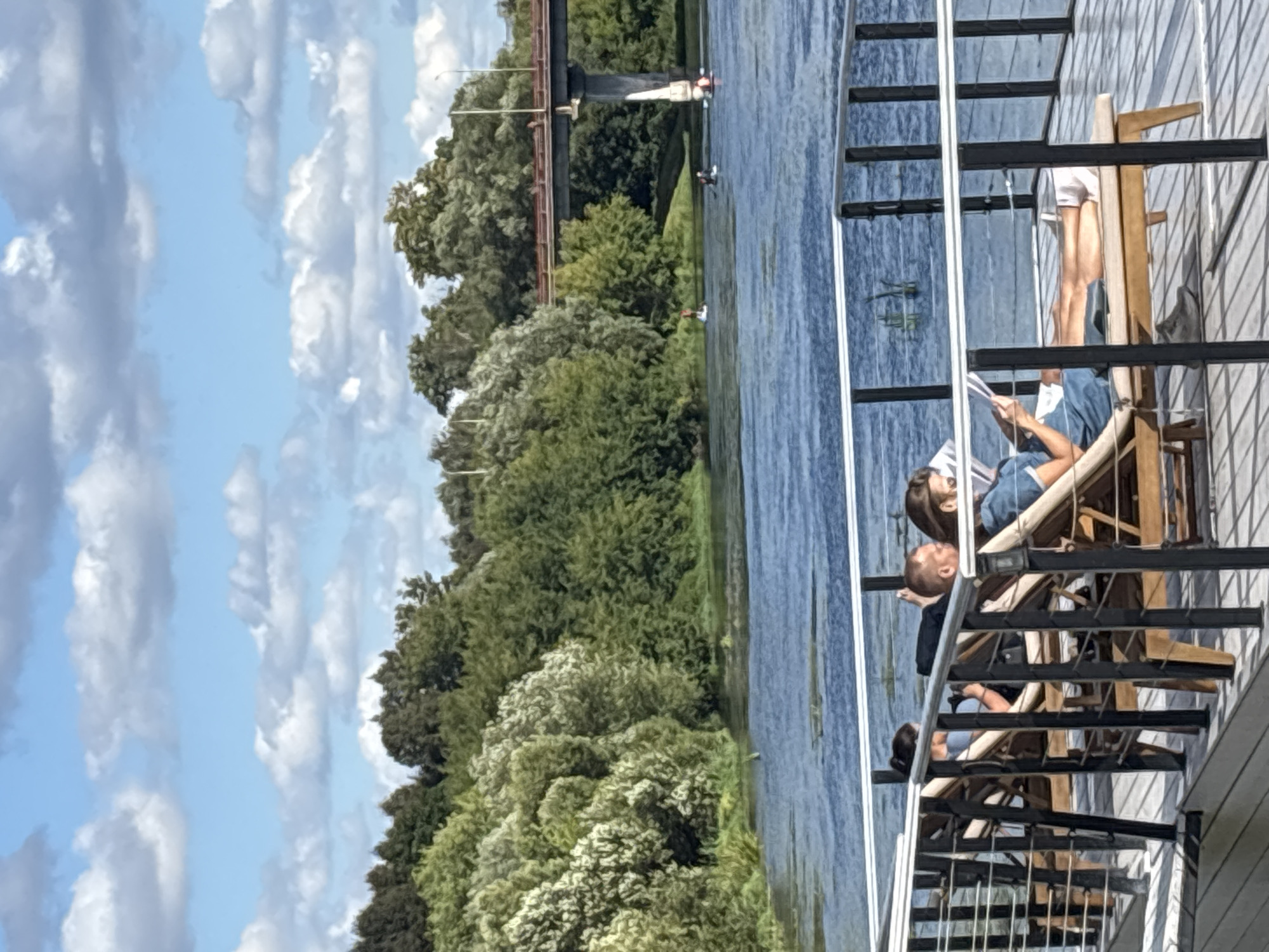 The image depicts a serene outdoor scene near the city of Zvenigorod, Russia, featuring a waterfront area with people relaxing and engaged in recreational activities. In the foreground, four individuals are seated on lounge chairs on a wooden deck, separated by black metal railings. One person, in the center right, is reading a magazine or book. To their left, a man is looking away from the camera, and further left, another person is partially visible. The deck overlooks a wide blue river, which has some scattered vegetation. In the middle ground, several people are on paddleboards in the river. Further in the distance, a large bridge with concrete pillars crosses the river, and beyond that, a dense line of green trees borders the opposite bank. The sky is a light blue with scattered white clouds, suggesting fair weather. The overall impression is one of peaceful leisure in a natural setting.
