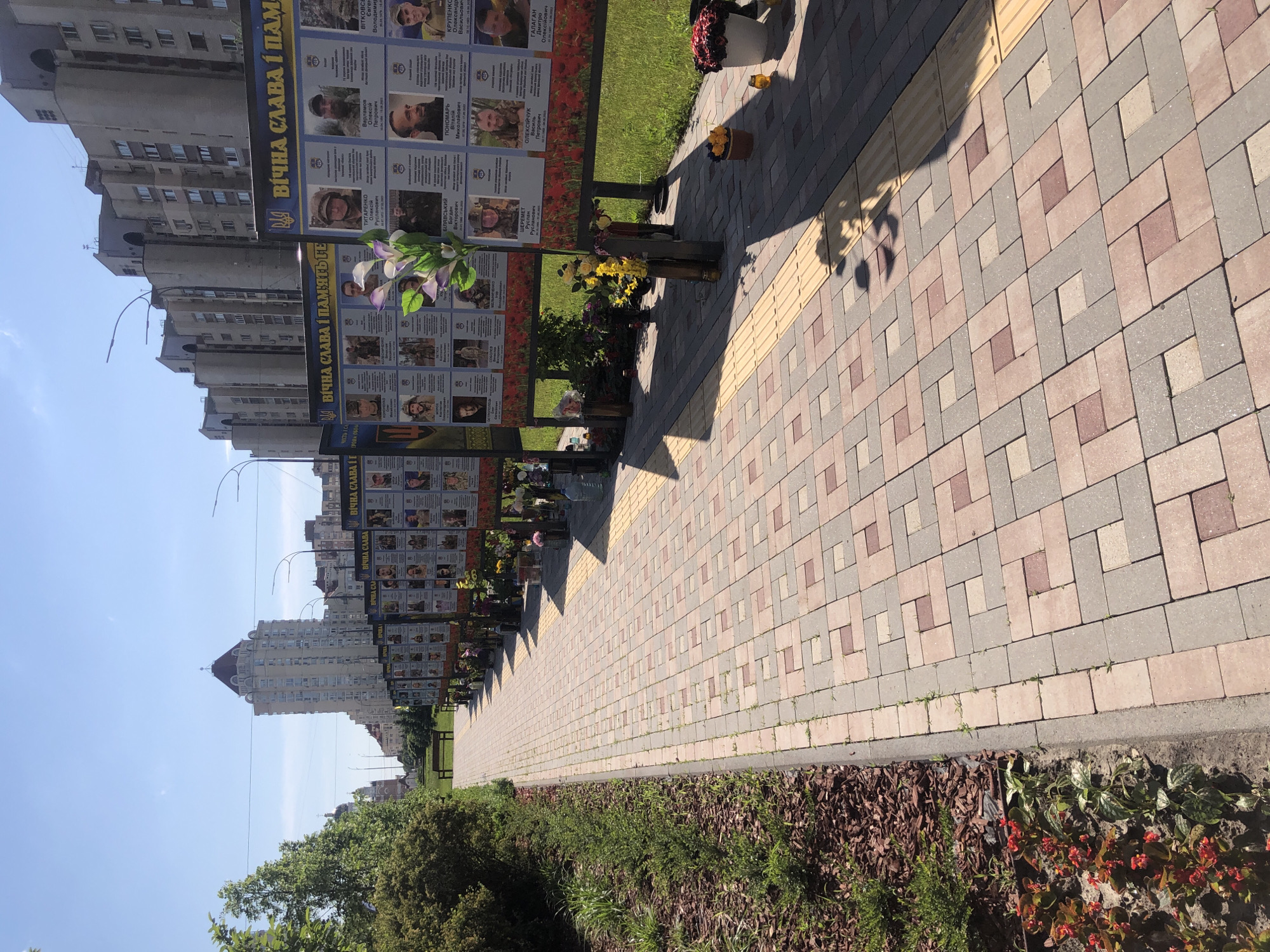The image shows a memorial walkway in Kyiv, Ukraine. Memorials are displayed along the walkway, containing portraits and names of fallen soldiers. The walkway is made of patterned brick and is lined with greenery. Buildings and a clear sky are visible in the background. There are several signs that say "Вічна слава пам'ять Героям" meaning "Eternal glory to the memory of the heroes" in Ukrainian.